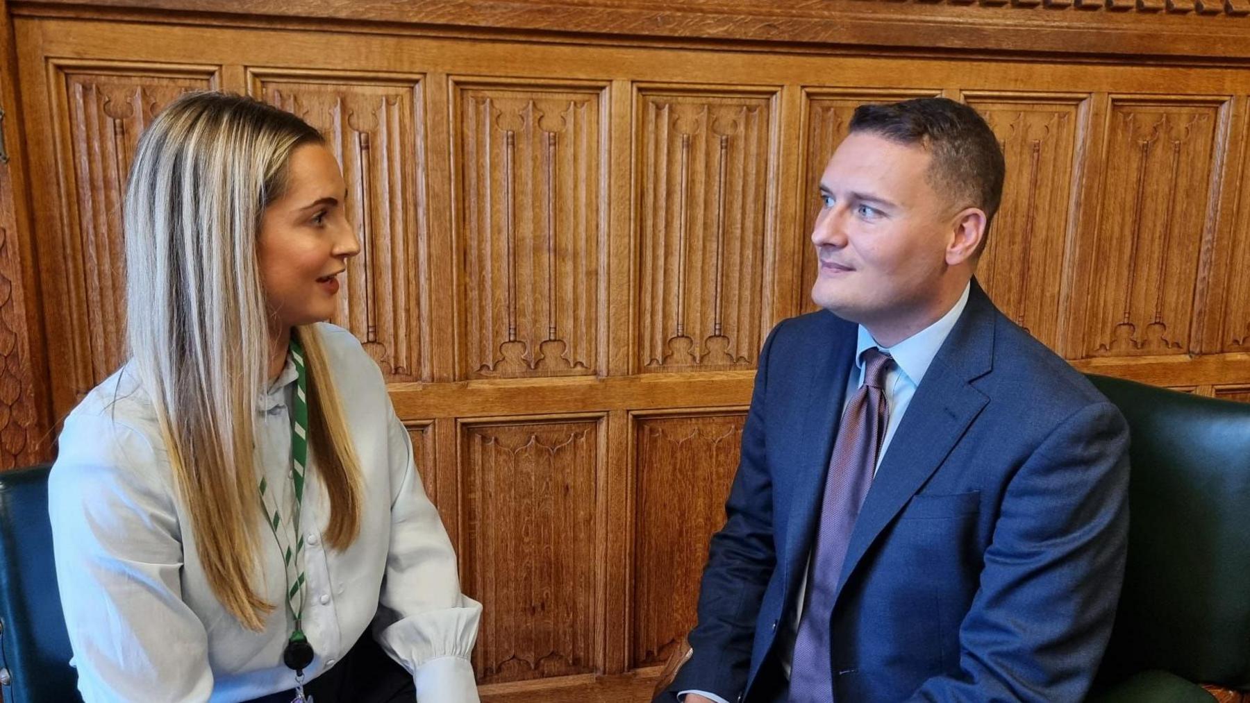 Rosie Wrighting with long blond hair wearing a pale blue top sits opposite Wes Streeting with short dark hair wearing a blue suit in front of wooden panelling.