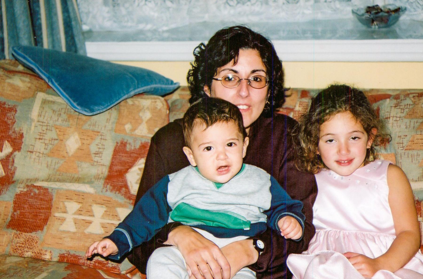 A family photo taken in the 90s of Sarah and her two children. They are sitting on a patterned sofa with autumn colours. Sarah has her toddler son on her lap and her daughter sitting next to her. 