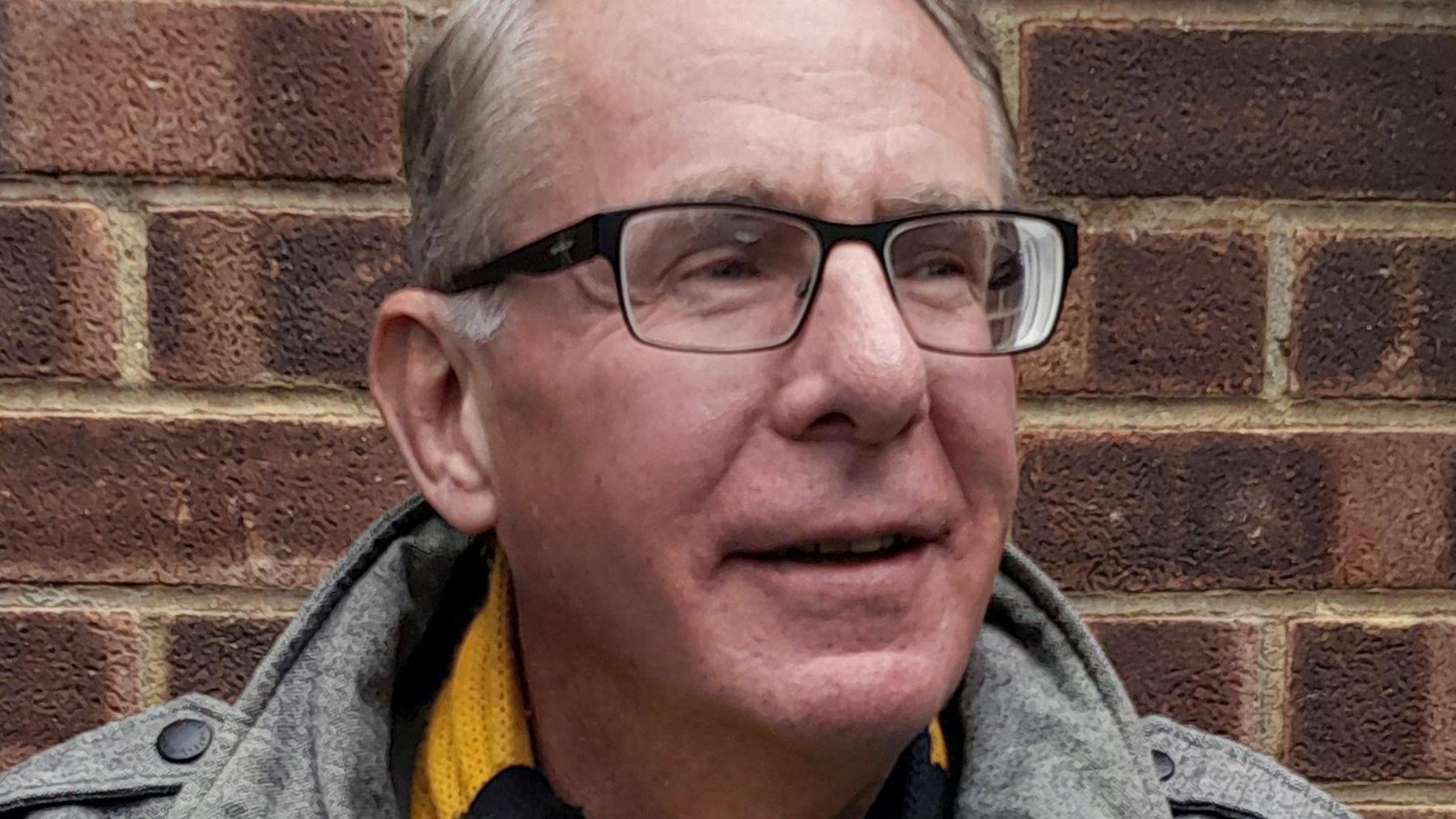 Nigel Pearce standing against a brick wall, wearing dark framed glasses, a grey casual jacket with the coller up and a yellow and black Cambridge United scarf, looking to his left