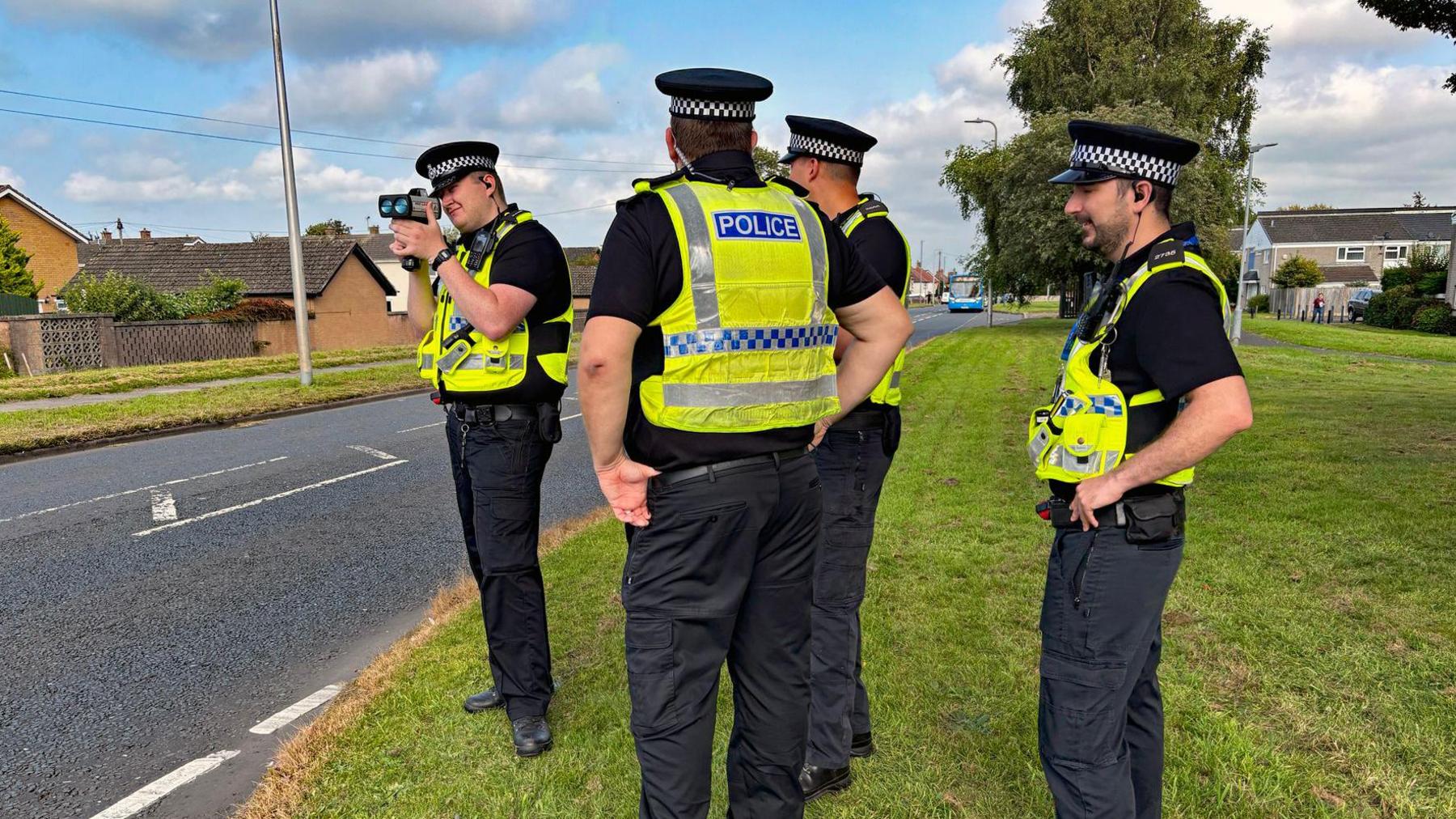 Four police officers in uniform standing on a grassy verge. One of them is squinting through a small device which looks like a speed camera.