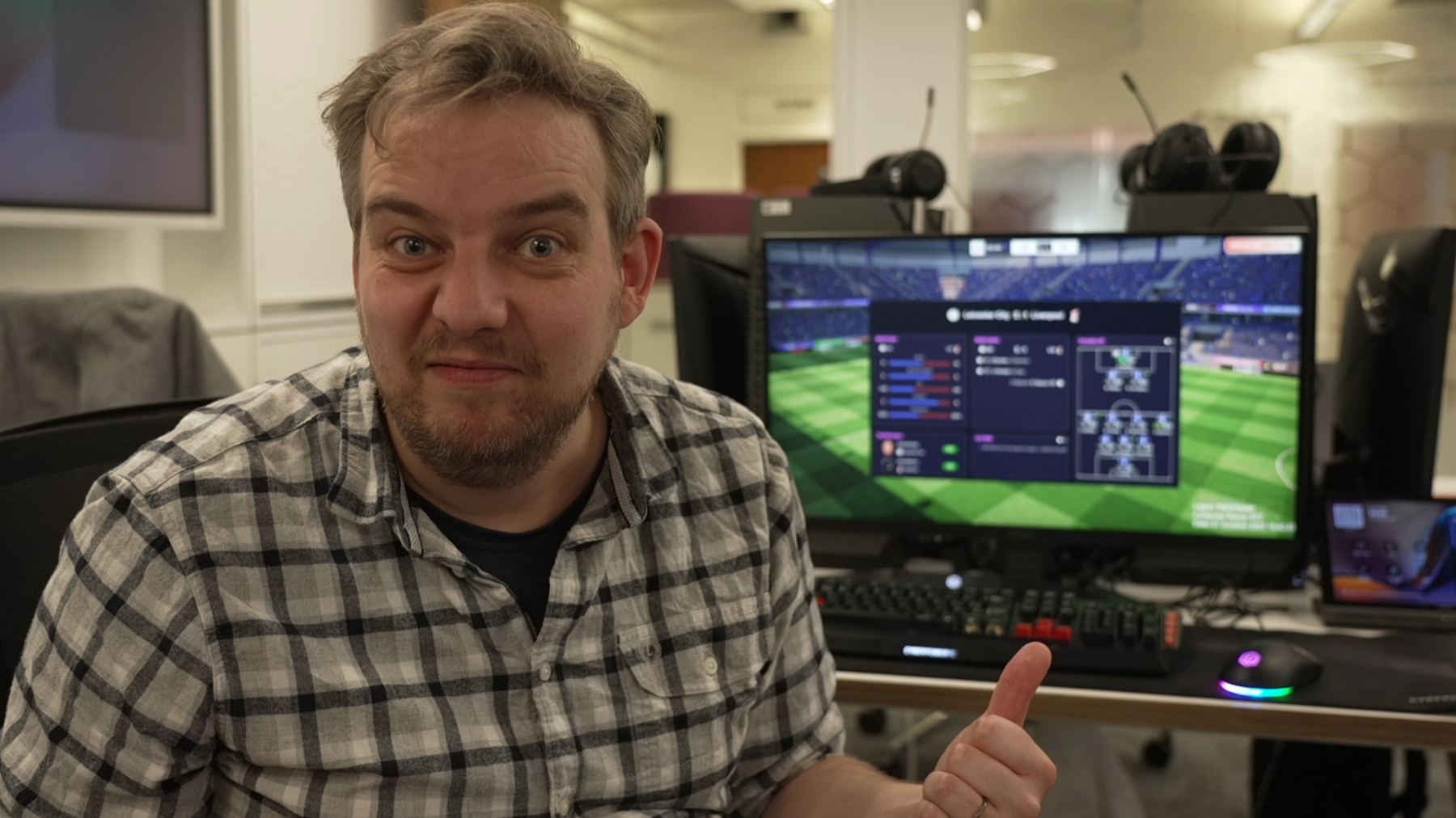 Tom Gerken sits at a computer. He is looking directly at the viewer and gesturing to the computer behind him, on which appears to be a video game screen.