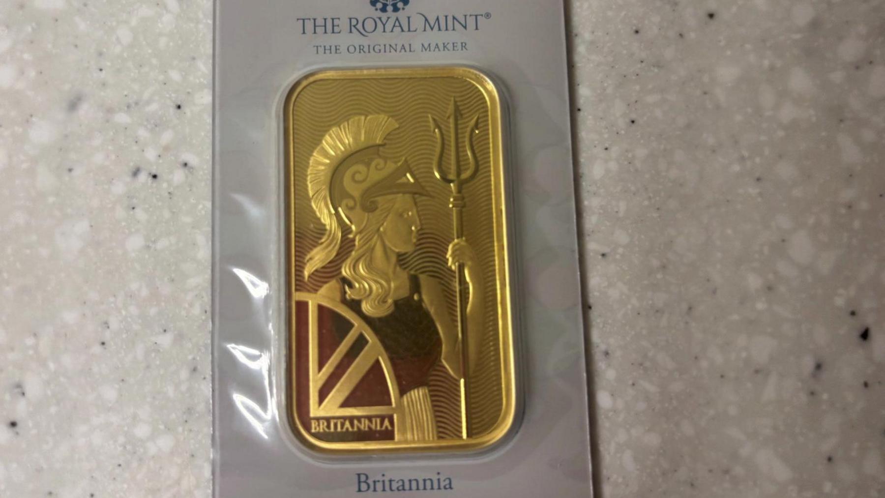 A gold bar from the Royal Mint sealed in packaging. The bar is solid gold with a soldier holding a shield and a spear.