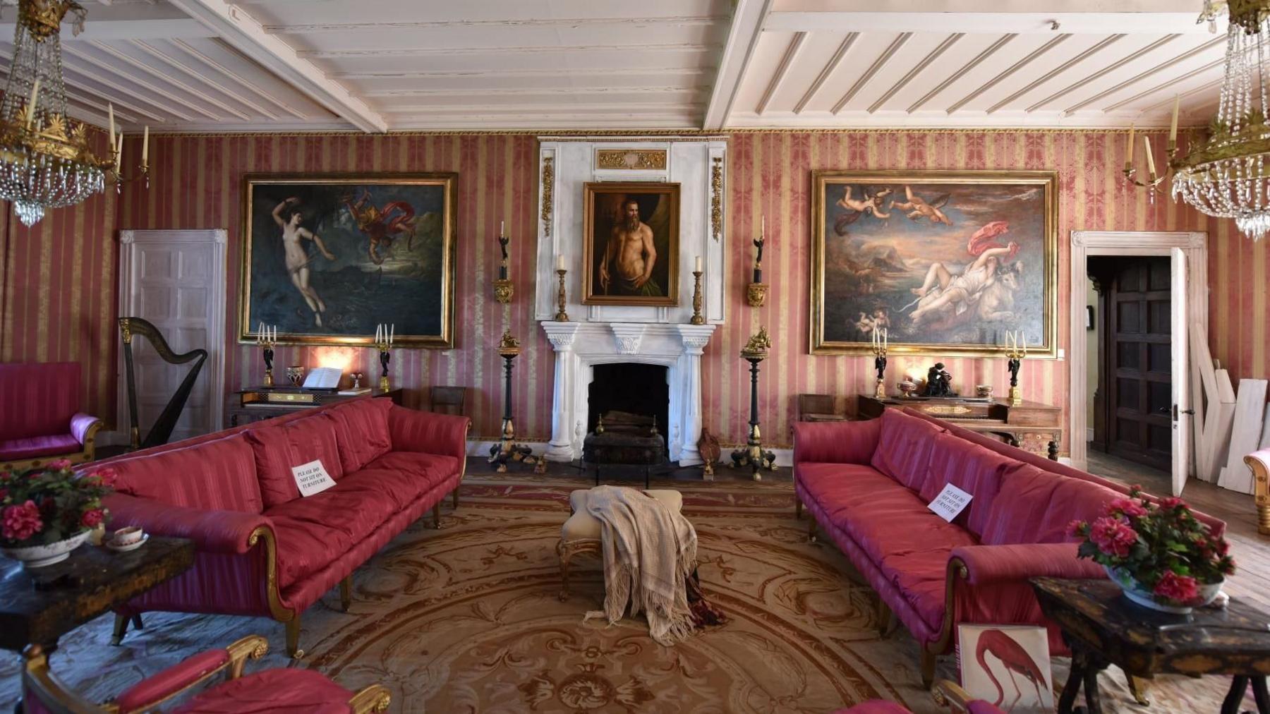 Sitting room with two red sofas, hanging chandeliers and numerous paintings on the walls