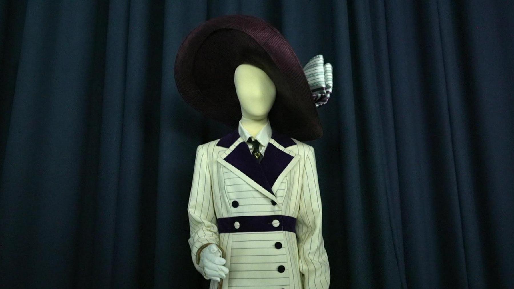 Kate Winslet's outfit on a mannequin in front of a dark curtained backdrop. it feature a wide-brimmed maroon hat with striped ribbons on the side. The white jacket dress is inspired by maritime design with blue pinstripes, buttons and trimmings. It features a shirt and tie beneath.