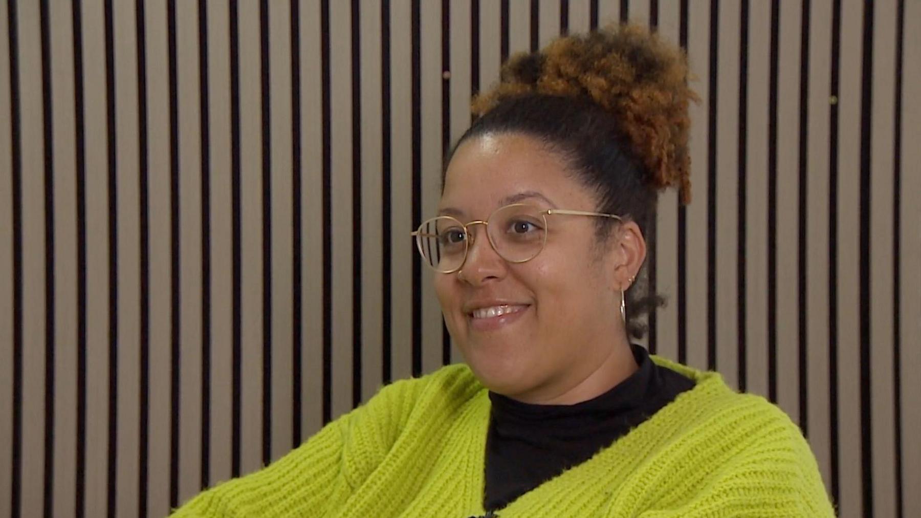 Kimberley is smiling. She is wearing a green jumper with a black t-shirt underneath. She has her hair tied up as bun on top of her hair and is wearing glasses.