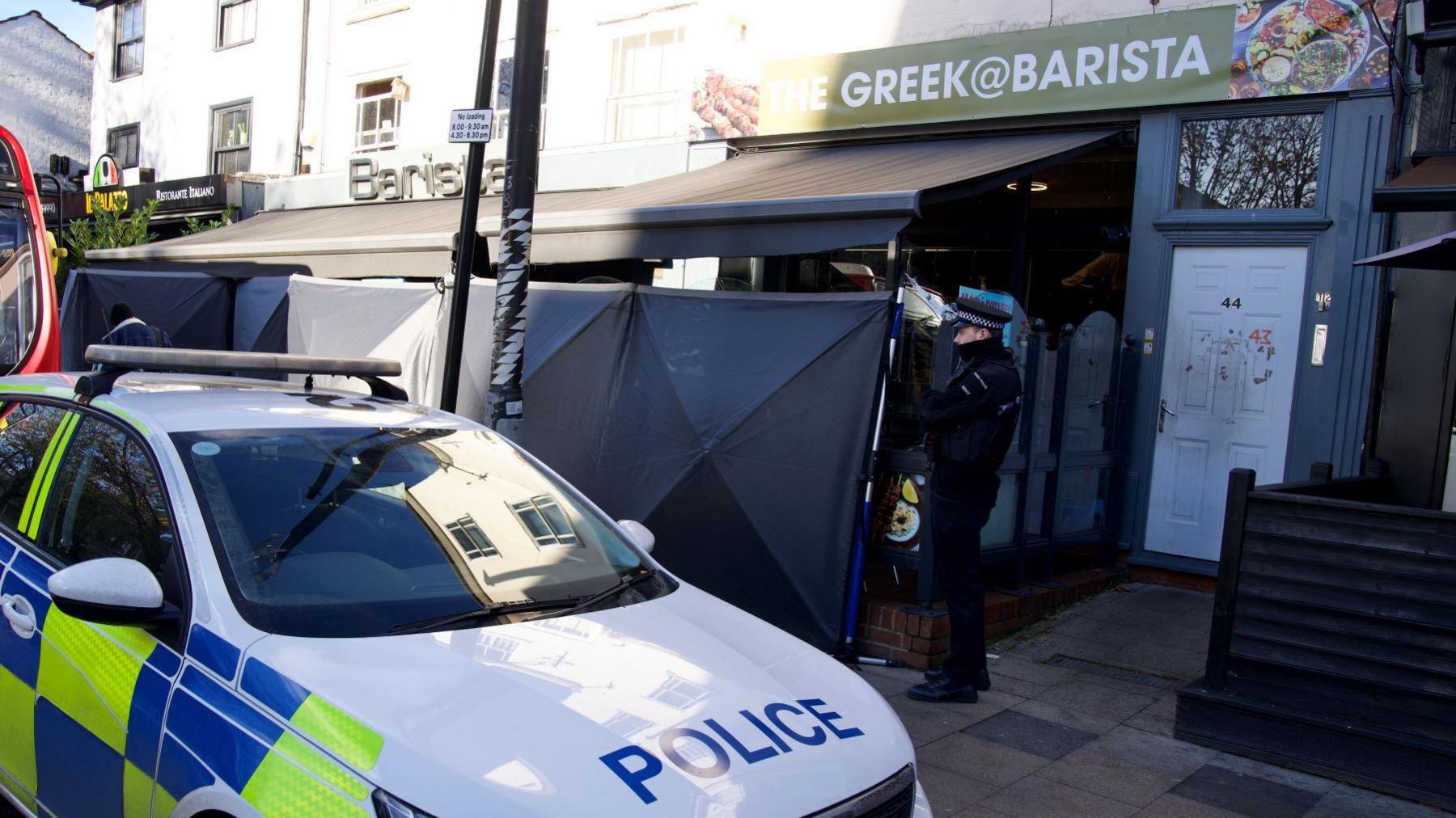 A police officer guards Barista bar in Chelmsford. The bar has black privacy screens around it and a police car parked out the front. The bar is set inside a terraced parade of shops.