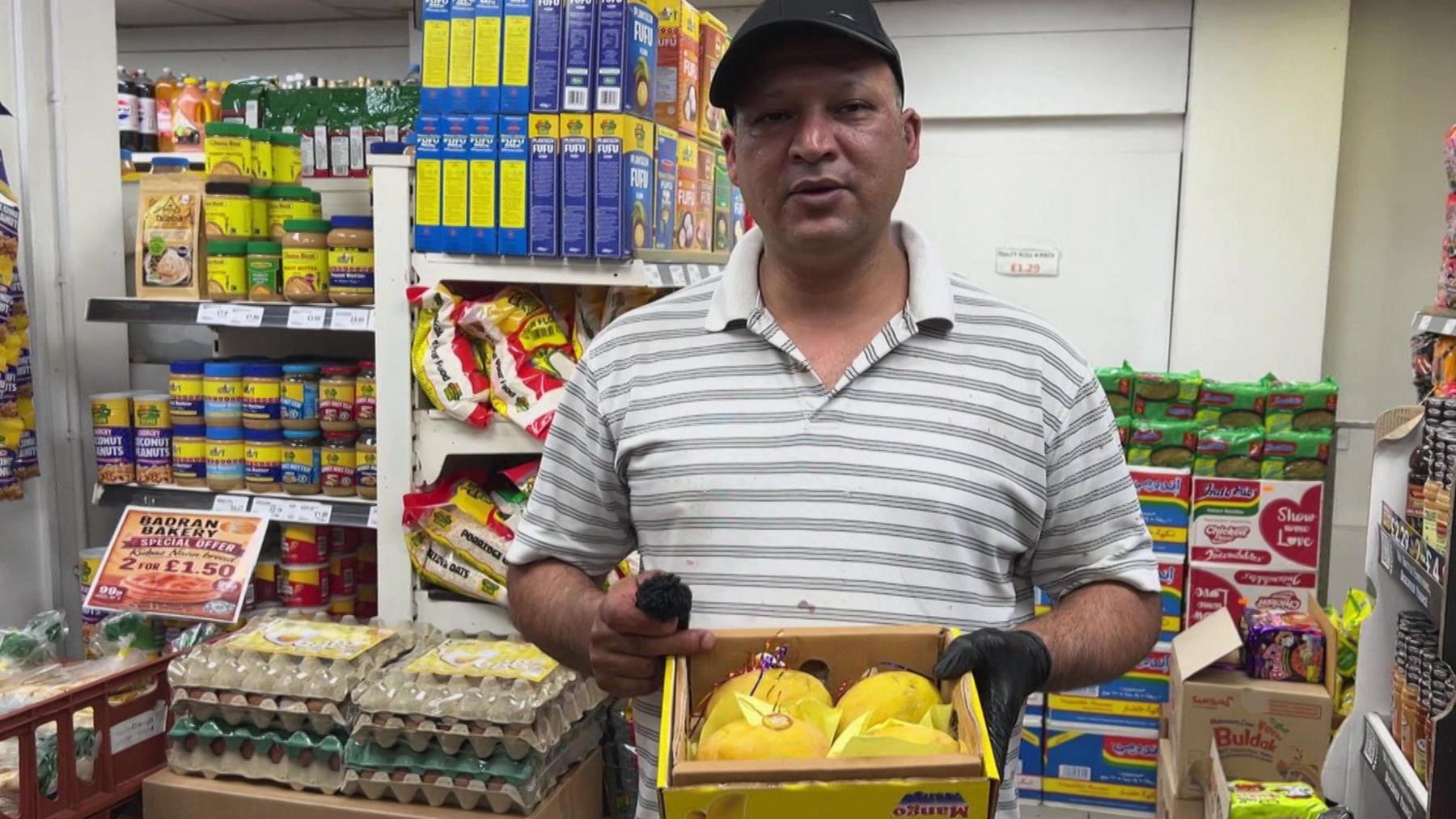 A man stands in the middle of a shop , surrounded by shelves of foodstuffs. He is holding a box of ripe, yellow mangoes. He wears a black cap and a striped shirt.