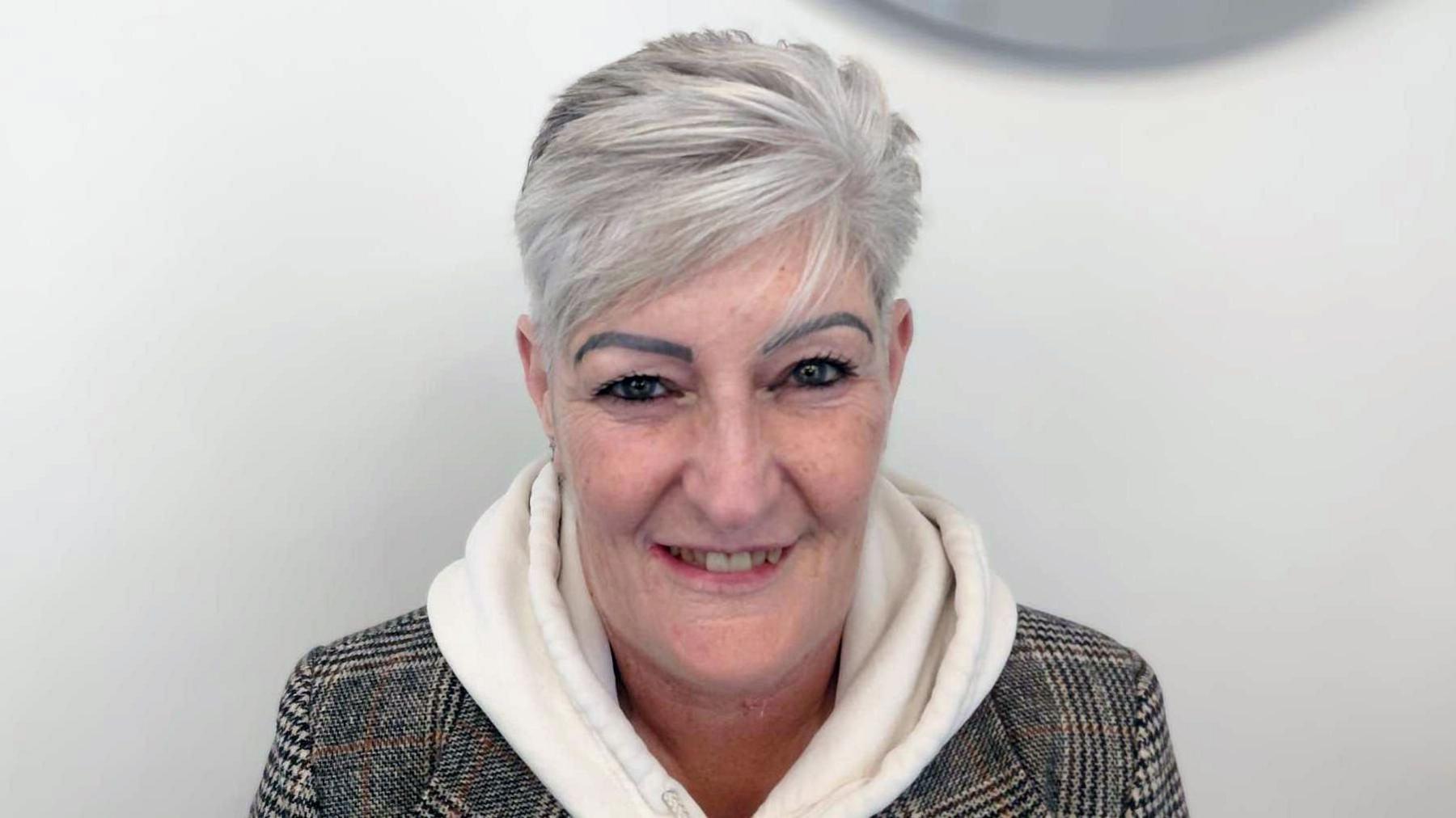 A head and shoulders crop of Val Lucus in a checked jacket and white hoody smiling and looking into the camera against a grey background.