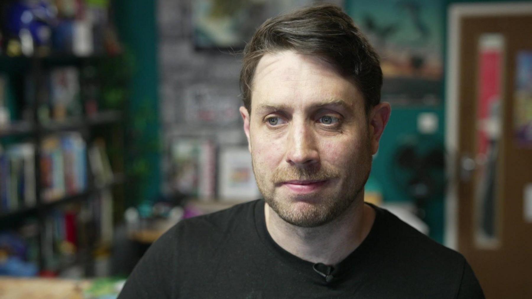 Nich Angell wearing a black t-shirt and sitting in his artist workshop. He has brown hair and a short beard and moustache. He is looking off to the left of the camera with a slightly concerned and despondent facial expression.