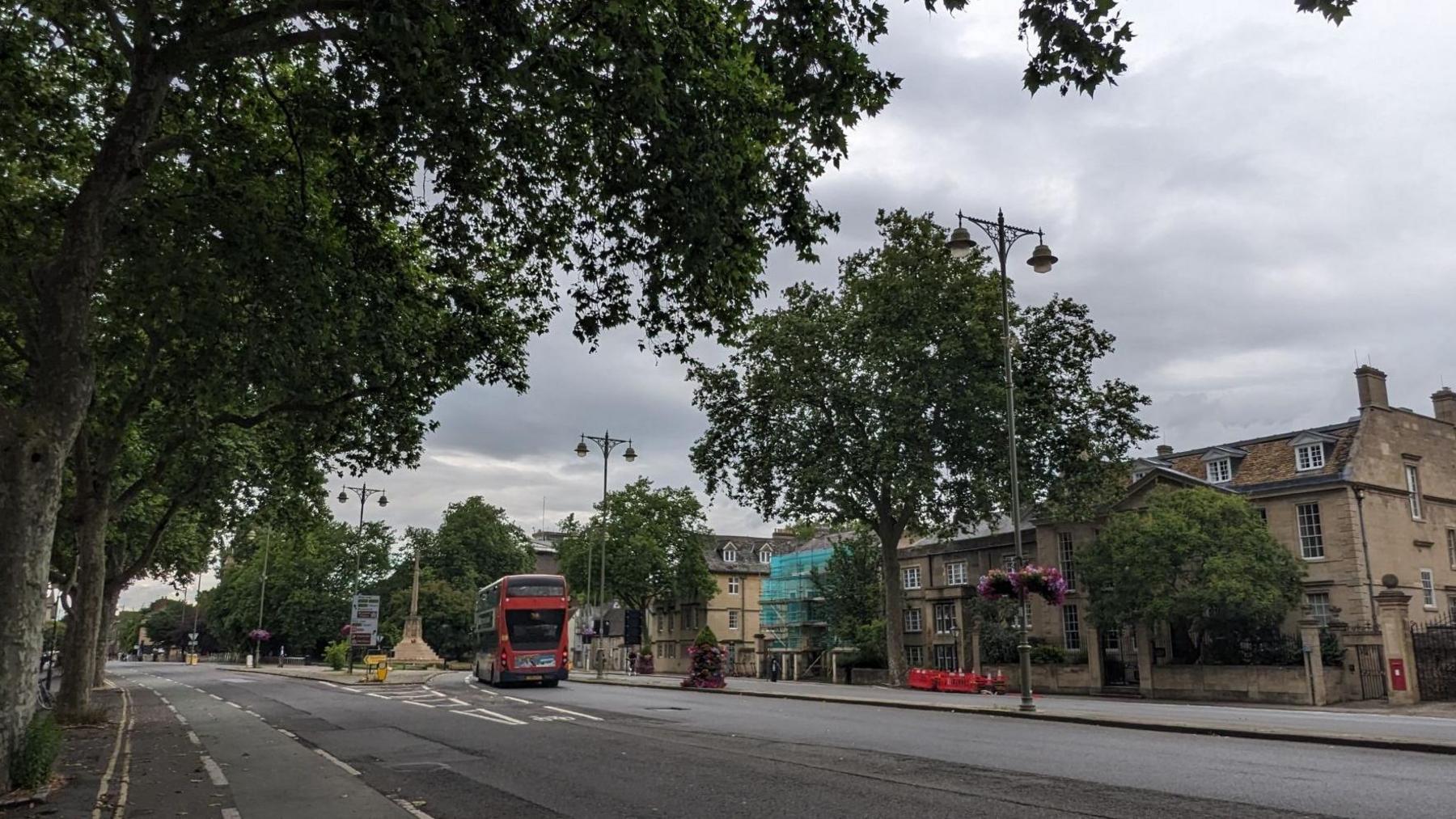 A road in a residential area. There is a red bus driving down the street. There are trees on the left side and buildings on the other. A cycle lane can also be seen.