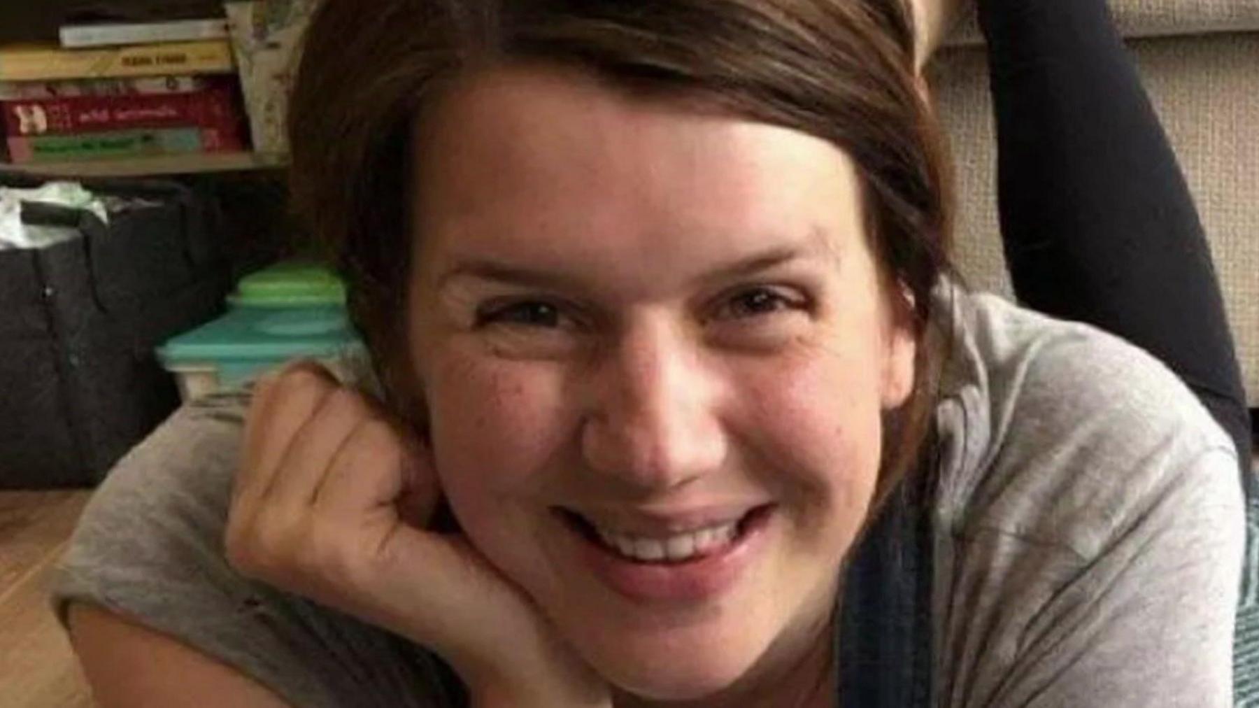Jennifer Cahill with long brown hair wearing a grey t-shirt in a home with books on a bookshelf behind her in the background. She is smiling.