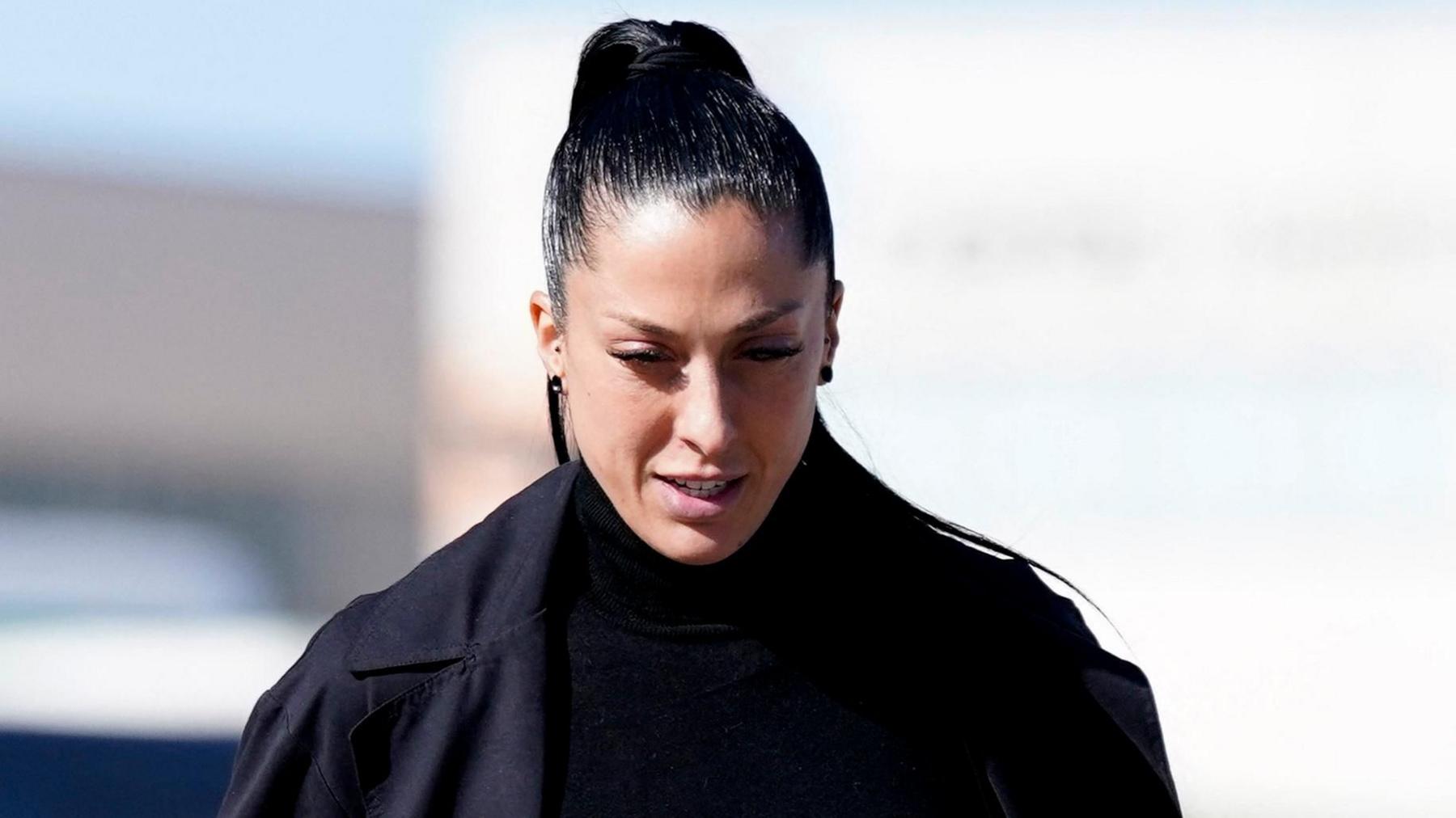 Jenni Hermoso looks down outside the court. She has long black hair slicked into a high pony tail and wears a double breasted black coat and black turtle neck