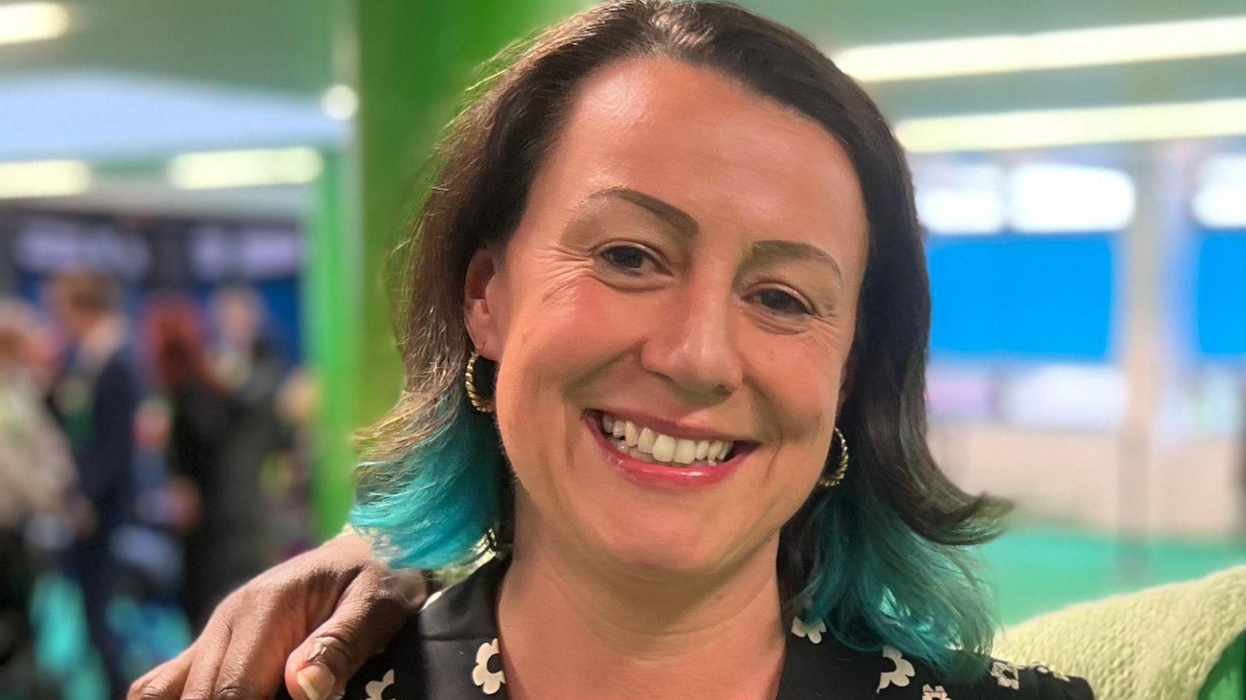 Emma Edwards smiling at the camera with dark hair that has blue highlights underneath wearing pink lipstick and earrings