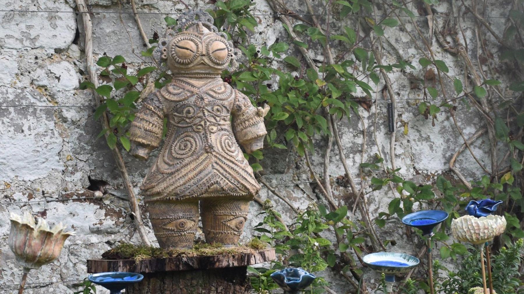 An ornate wood carving of a person featuring patterns in lines and circles over their body is called Ancient Protector by Maria Alvarez Echenique. 