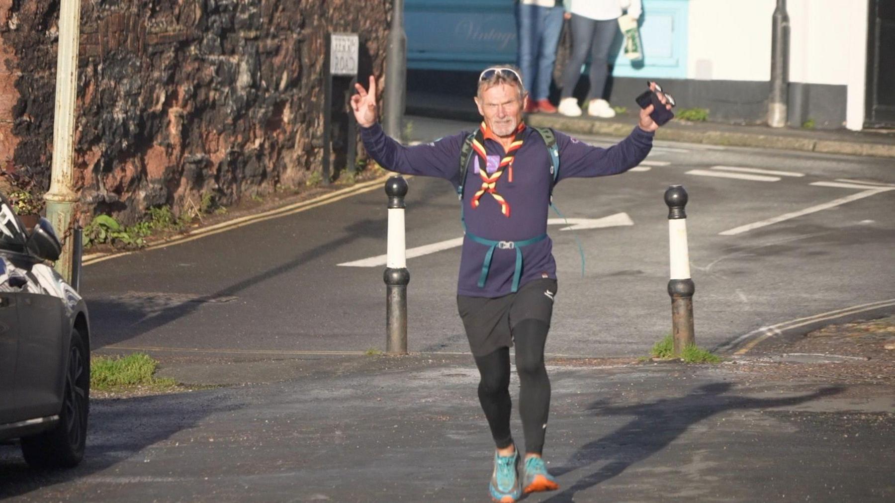 A man with silver hair running on a road with his hands in the air. He has a mobile phone in his hand and a pair of white-framed sunglasses on his head.