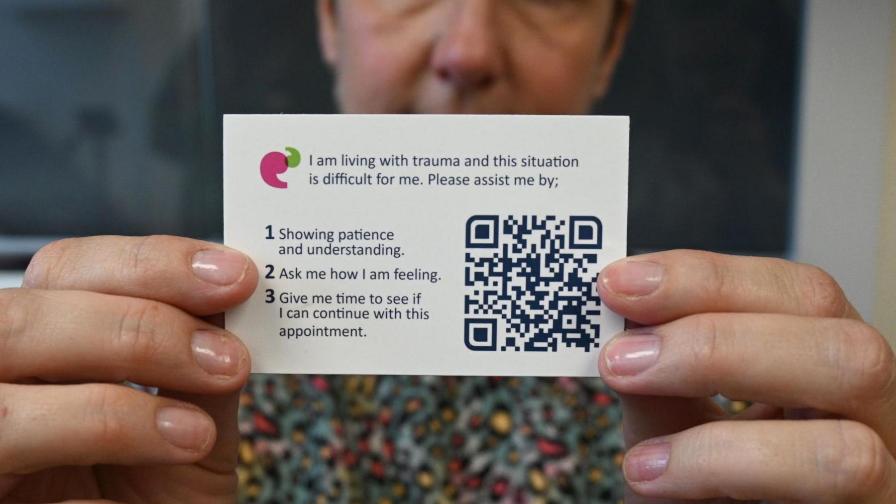 A woman, who is blurred, holding a white trauma card in front of her. The card advertises the cardholder is a trauma survivor. It alerts people to this fact and tells them how they can help.