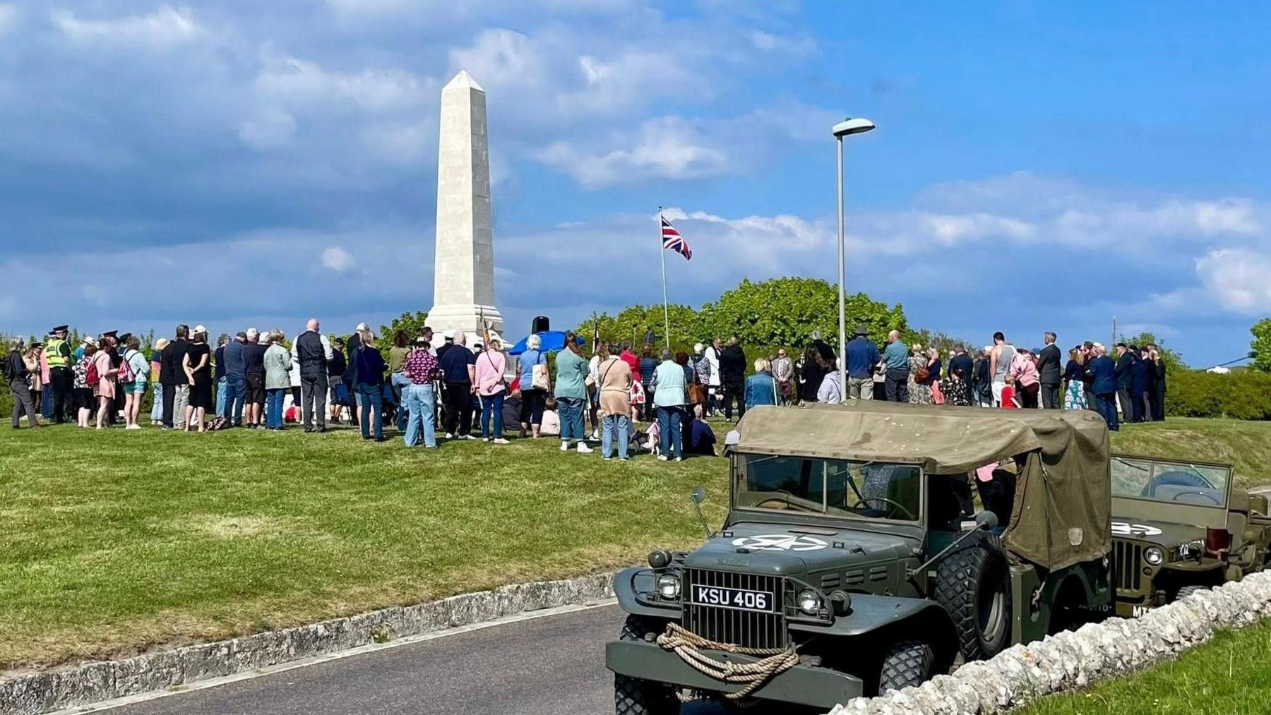 A large group of people gathered around a war memorial on an area of grass. In the foreground is a road and two parked world war two-era army vehicles. It's a sunny day and the sky is blue with scattered clouds.