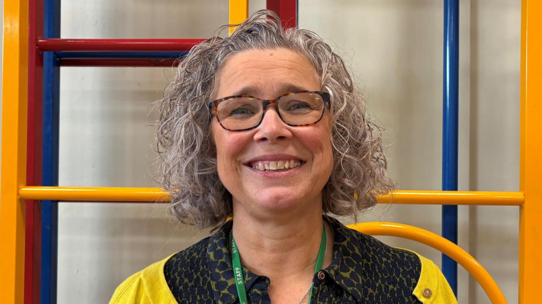 A woman from the chest up is smiling directly at the camera while in front of a red and orange climbing frame. She is wearing a yellow cardigan and a black spotty dress. She has glasses and curly, bob length hair.