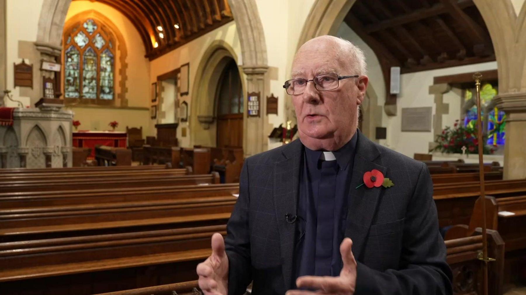 The Reverend Phil Hanson stands near the pews. He is wearing clerical attire and has a poppy on his lapel. The church pulpit and altar and a stained glass window can be seen in the background. 