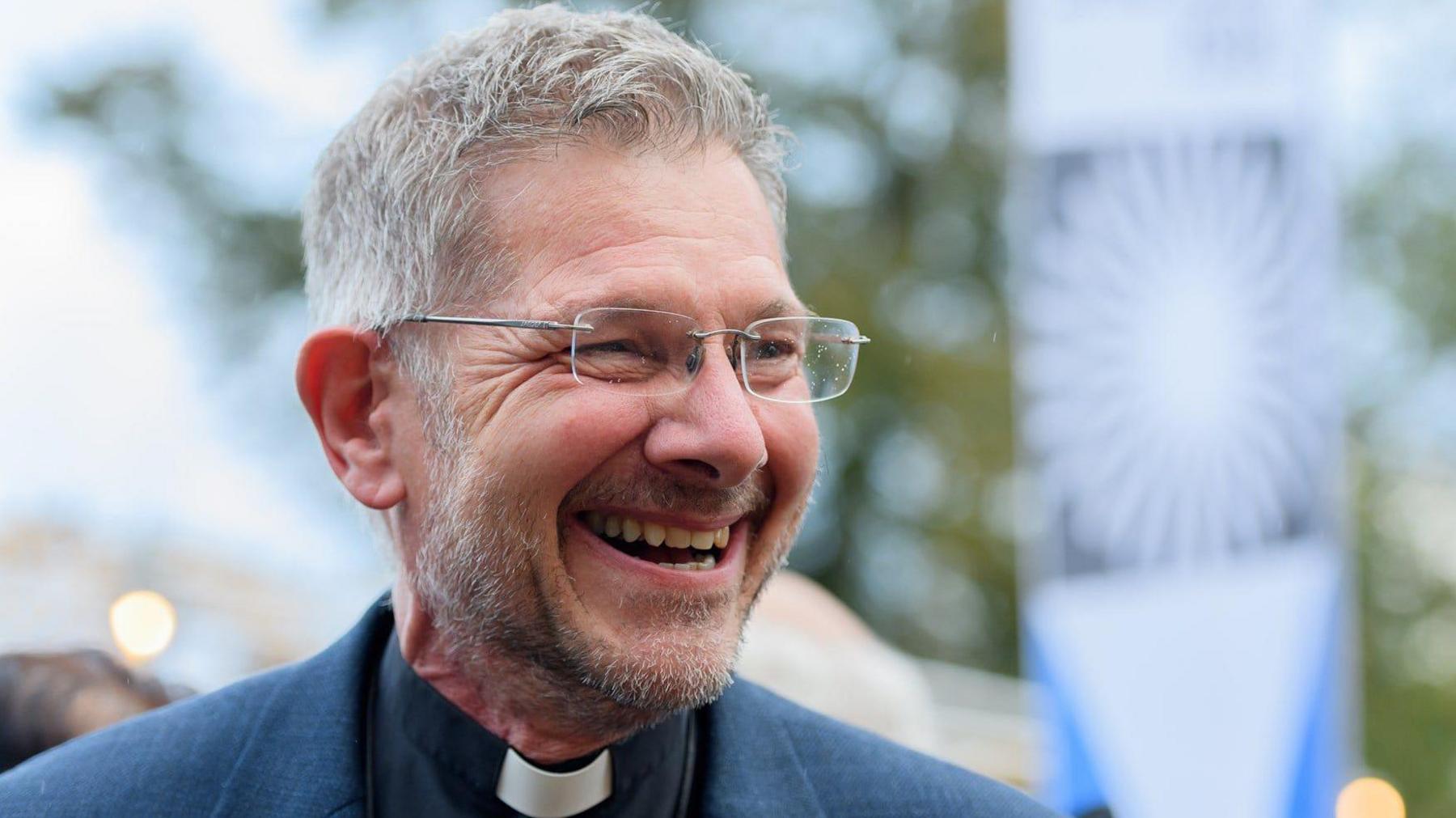 A man with short grey hair, glasses, a vicar's collar and a blue jacket, looks to his left and smiles.