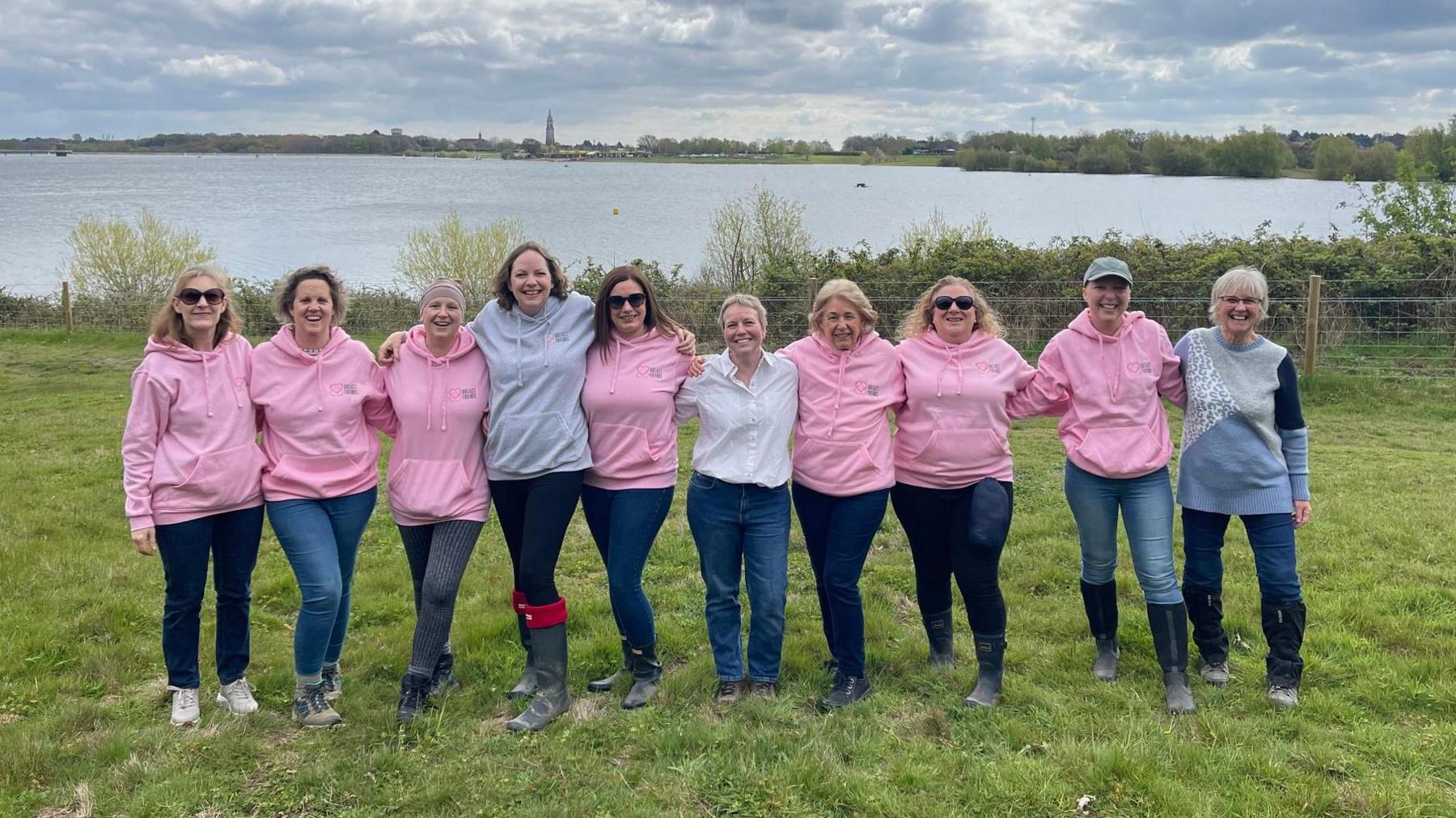 A row of woman – some wearing pink hoodies – standing on grass while smiling at the camera