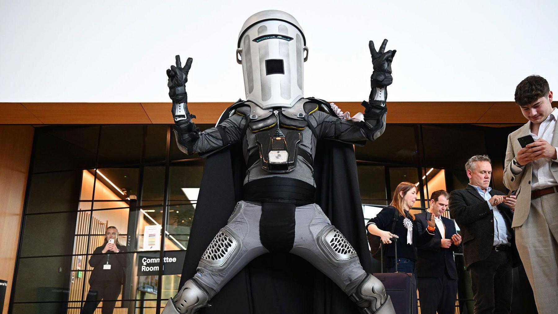A man with a bin on his head poses triumphantly outside a giant glass window. The bin is silver with a slit cut out for his eyes and a square hold for his mouth. He also wears black gloves, a black breastplate, knee and elbow guards, and a long silver cape. 
