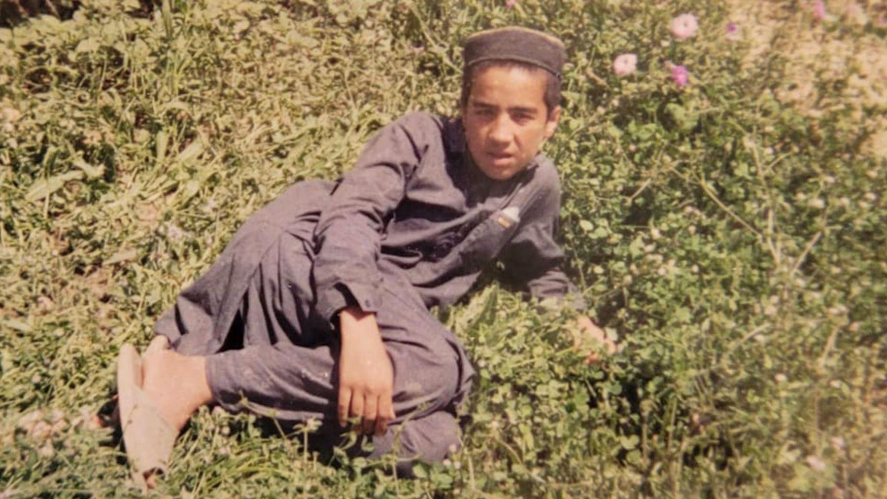 A colour photo of a teenage boy lying on his side on a patch of ground that is covered in green foliage. He is wearing a traditional dark blue Afghan tunic, trousers, sandals and brimless hat.