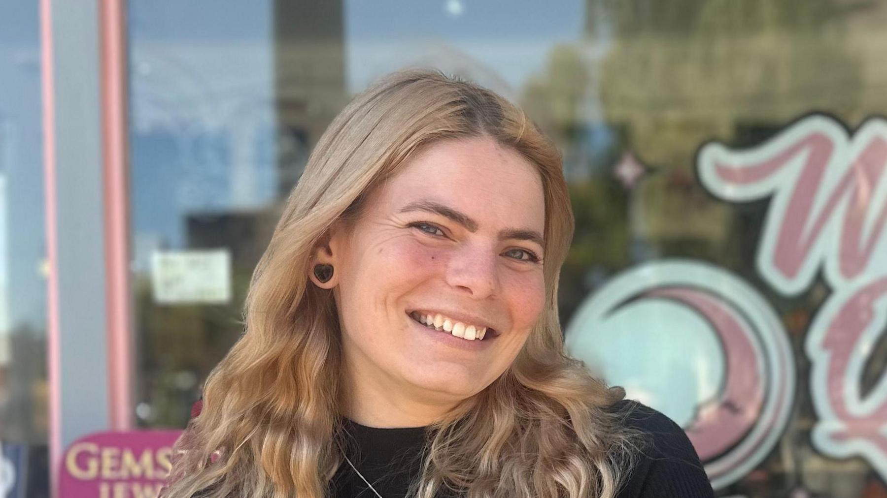 Lawra Stubbs smiles at the camera outside her shop. She has long blonde hair that is slightly crimped. She has a large black earring in her left ear and wears a black top.