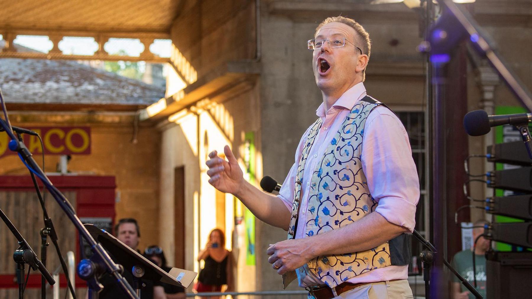 A man conducting a choir. He has short hair and is wearing reading glasses, a baby pink shirt, beige trousers and a beige and blue patterned waistcoat. His mouth is open and one of his hands is raised in line with his chest. He is not looking at the camera.
