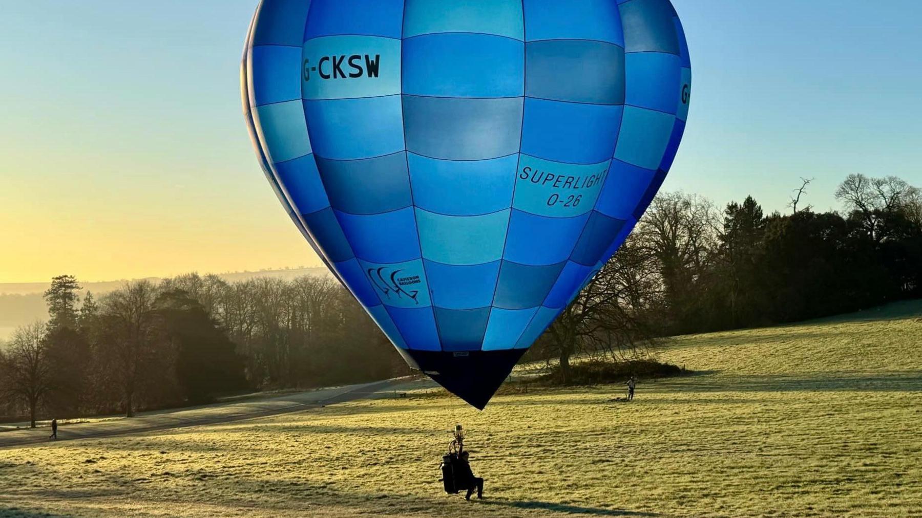 A man sits in a small seat underneath a medium-sized hot air balloon close to the ground in the parkland of Ashton Court near Bristol. It is a sunny day, with the picture taken not long after dawn. The balloon is called a Cameron Superlight O-26 G-CKSW