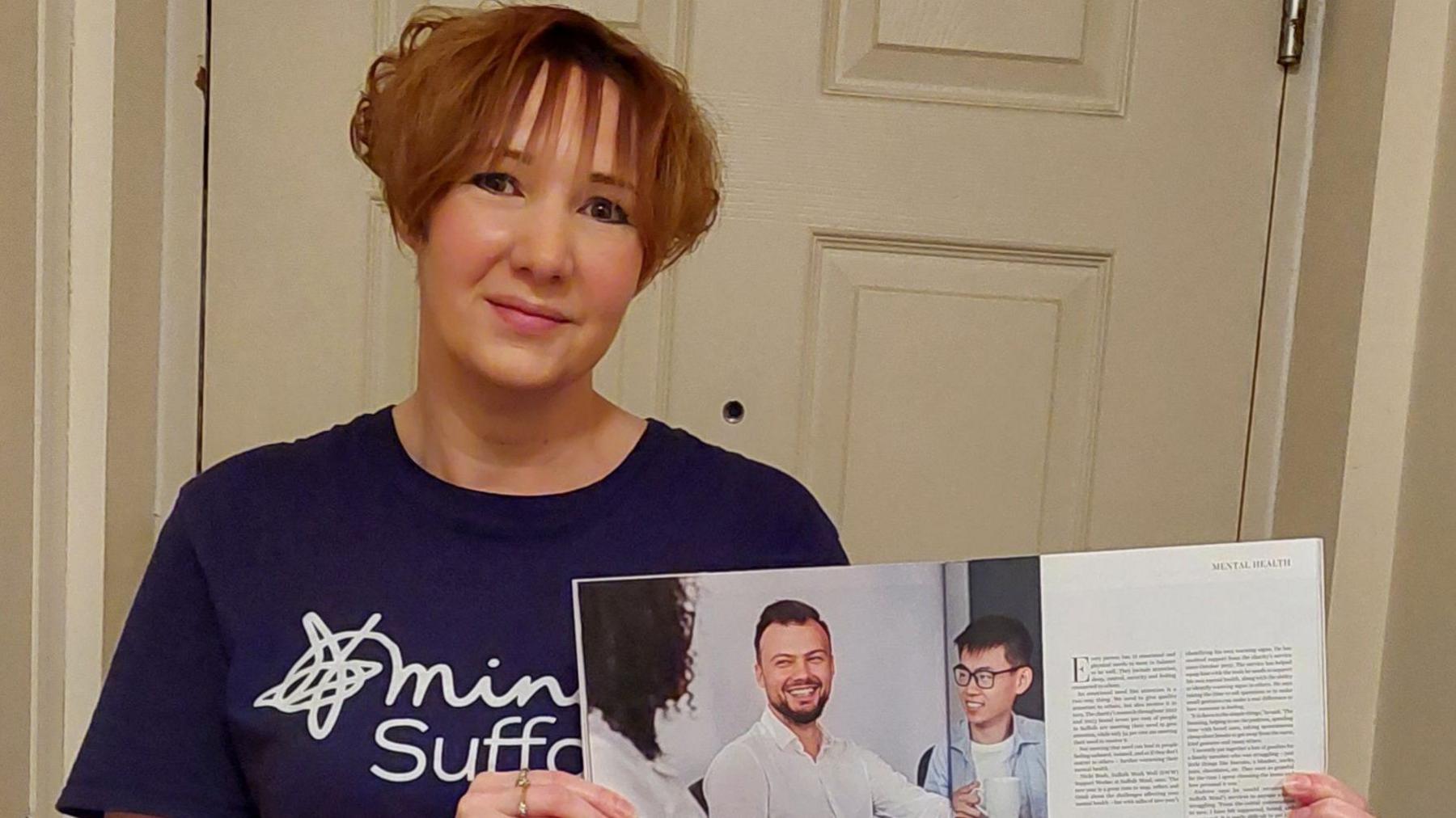 Nicki Bush - a woman wearing a blue Suffolk Mind t-shirt while standing in front of a door. She is holding a magazine. 