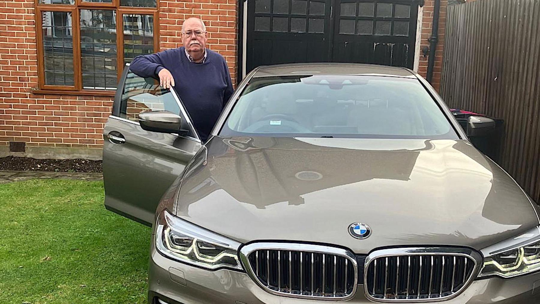 Chris stands beside his grey BMW 5 Series parked on a driveway outside a red-brick house, with one hand resting on the open driver’s door.