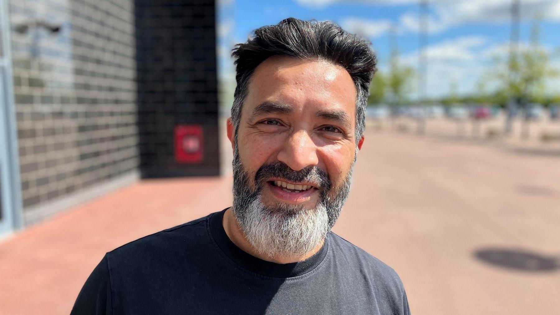 Man with grey t shirt and beard standing outside Stadium MK