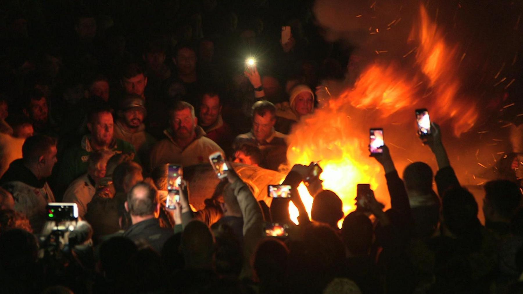 A group of people with their arms in the air film a flaming barrel on their phones. It is dark.