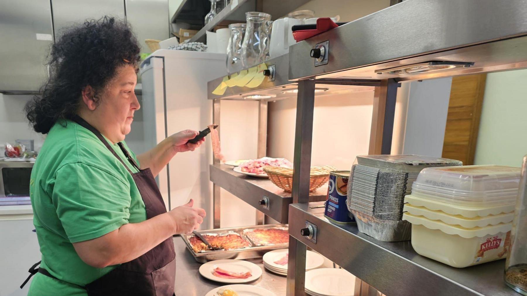 The picture shows a person in a kitchen setting, wearing a green shirt and a black apron, preparing food. The individual is working at a counter with a variety of fresh ingredients and cooked dishes, including sliced vegetables, lettuce leaves, and trays of prepared food. There are also containers of ingredients like eggs and other packaged items on the counter. The background features kitchen equipment, shelves with glass jars, and utensils.