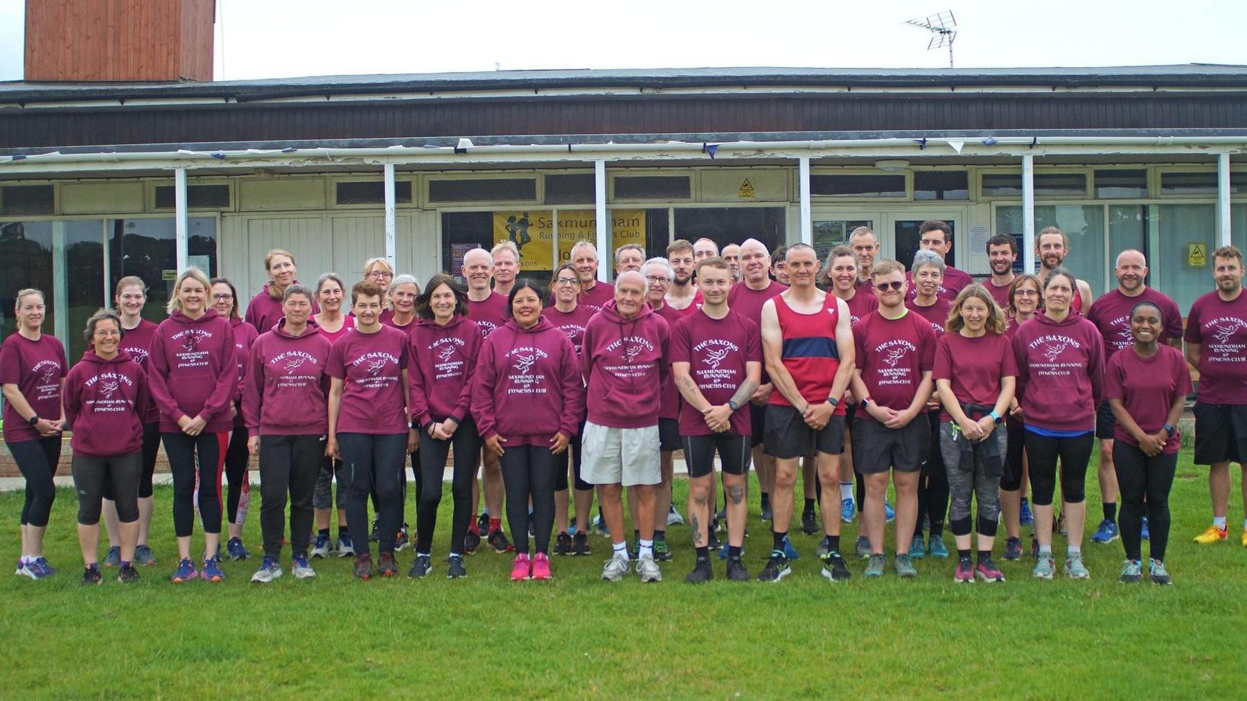 A large group of people stand on a grass sports field outside a pavilion. They all wear matching dark purple hoodies with the running clubs logo in white on the front. They smile at the camera.