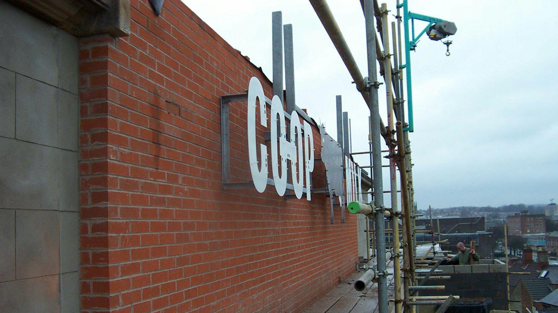 A photograph from the side of a large sign saying "Co-op Milk" with a cow in the middle. The sign is on a brick building and it is daylight. 