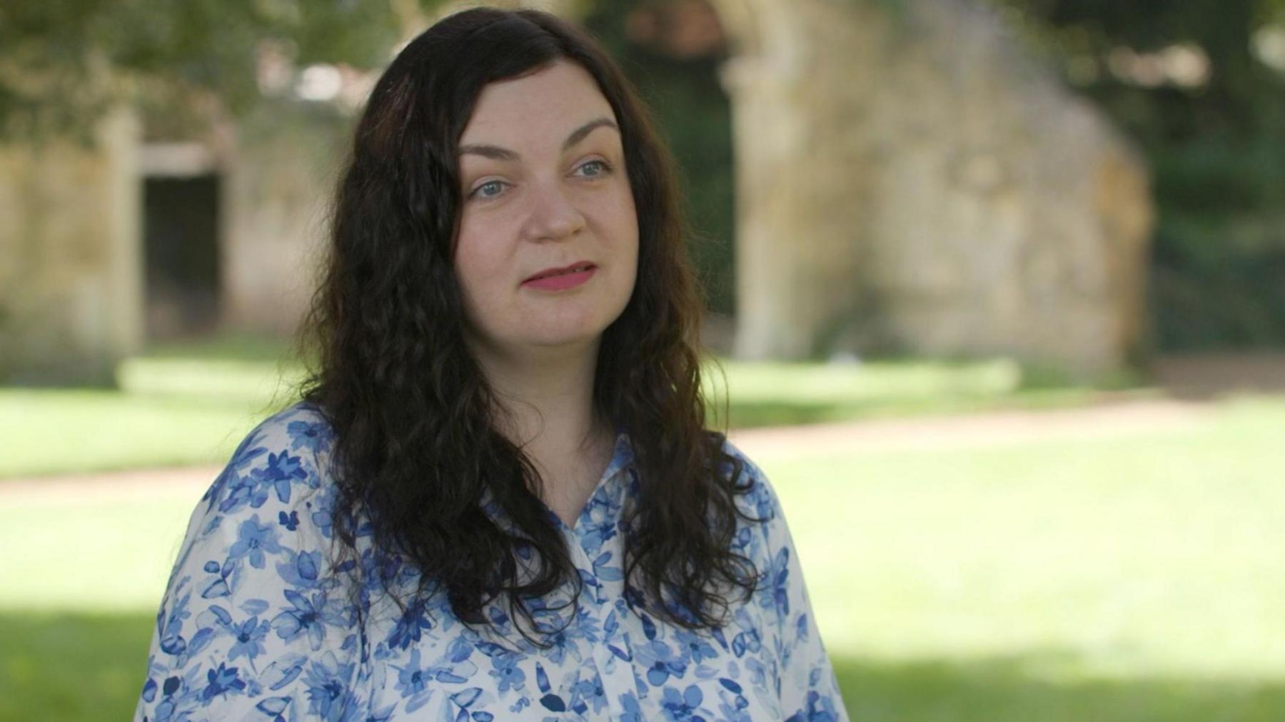 Olha looks slighty off camera during her interview, she has mid length wavy black hair and wears a white dress with a pale blue floral print. The greenery and building is in soft focus behind her.