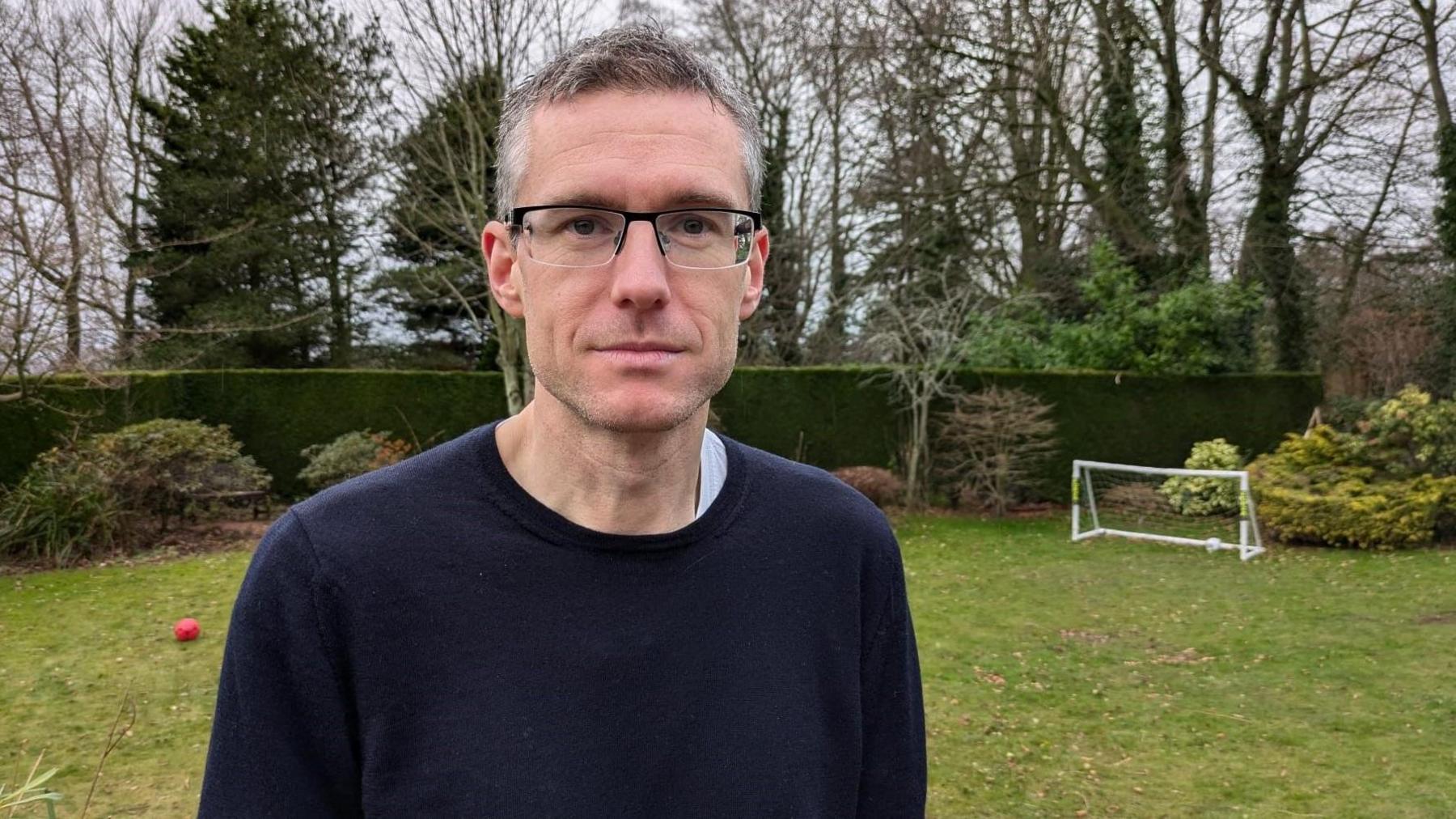 Mark Church is standing in his garden. He is in his 40s and wearing a blue jumper. Behind him is a hedge and some miniature football goalposts 