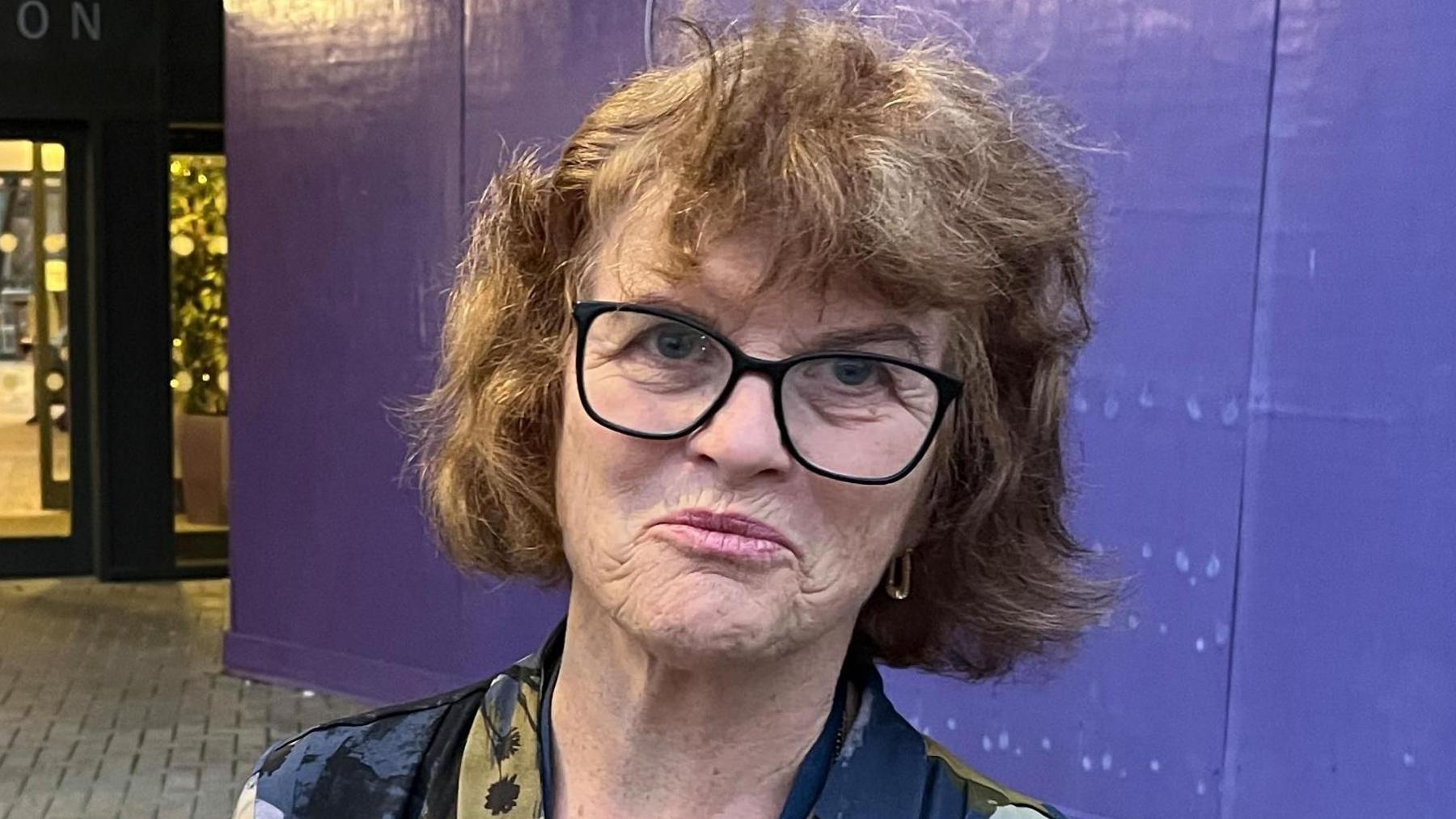 A woman with glasses in front of a building entrance which has large glass doors and a sign above that reads "PRIORY VIEW RECEPTION". There is a pavement area with a purple wooden barrier on the right. The woman is wearing a patterned top with shades of blue, green, and grey, and a necklace. There is a reception area visible through the glass doors.