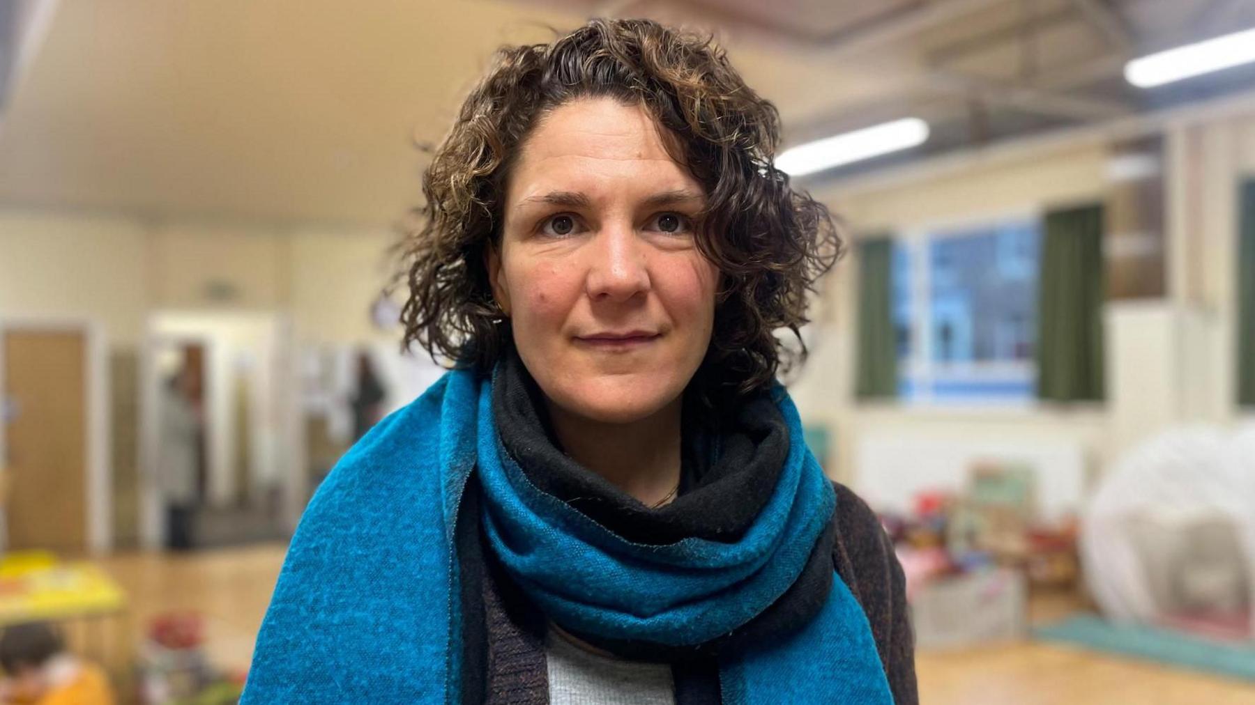 A woman with dark curly hair and wearing a thick winter scarf smiles at the camera. A blurred nursery room is in the background. 