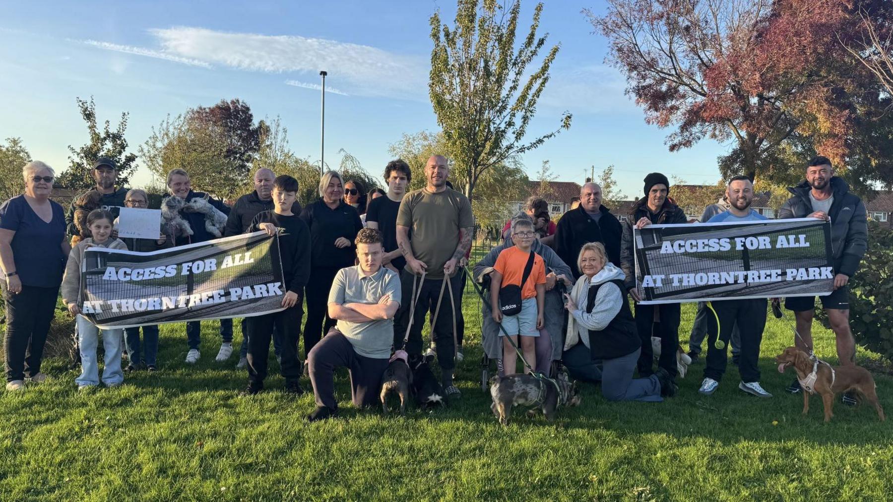 About two dozen people are standing in a park and facing the camera, with some pulling defiant poses. They are holding up signs that say: "Access for all at Thorntree Park".