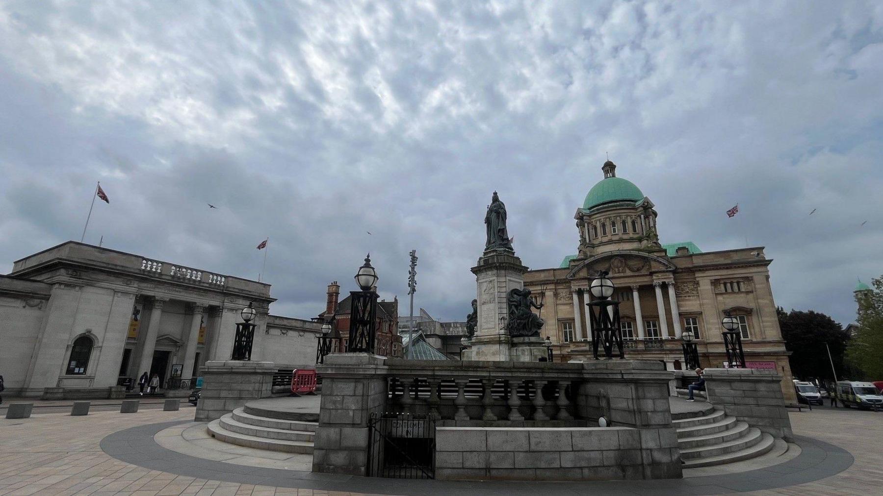 Queen Victoria Square in Hull. A statue on a plinth is at the centre of the image and surrounded by large stone steps. The Ferens Art Gallery is visible on the left side of the image, and City Hall, with a green copper dome on its roof, is in the background.