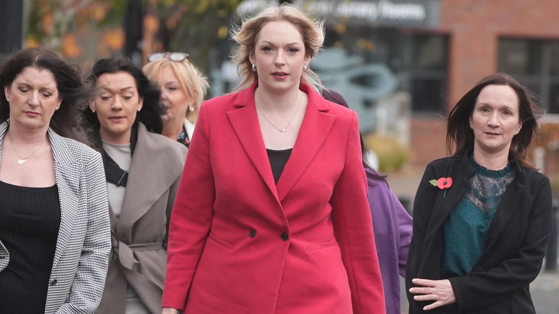 A group of women arrive at the tribunal office. At the head of the group is Bethany Hutchison, who has long blond hair and is wearing a red coat. Beside her are several smartly dressed women with long brown hair.