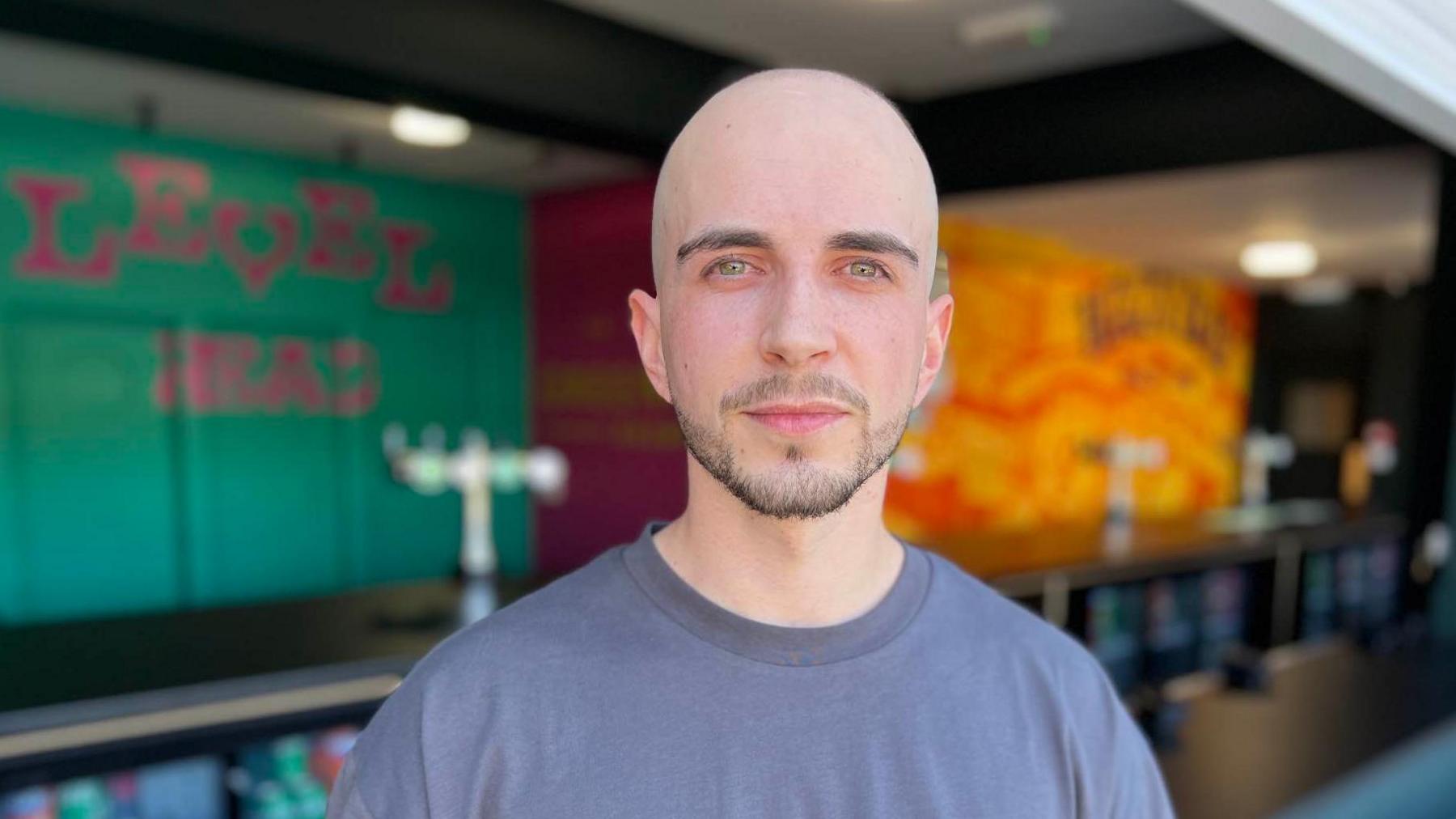 Madalin Acunune who has shaved head and a close-cropped dark beard wearing a dark t-shirt looking towards camera while standing in front of a bar