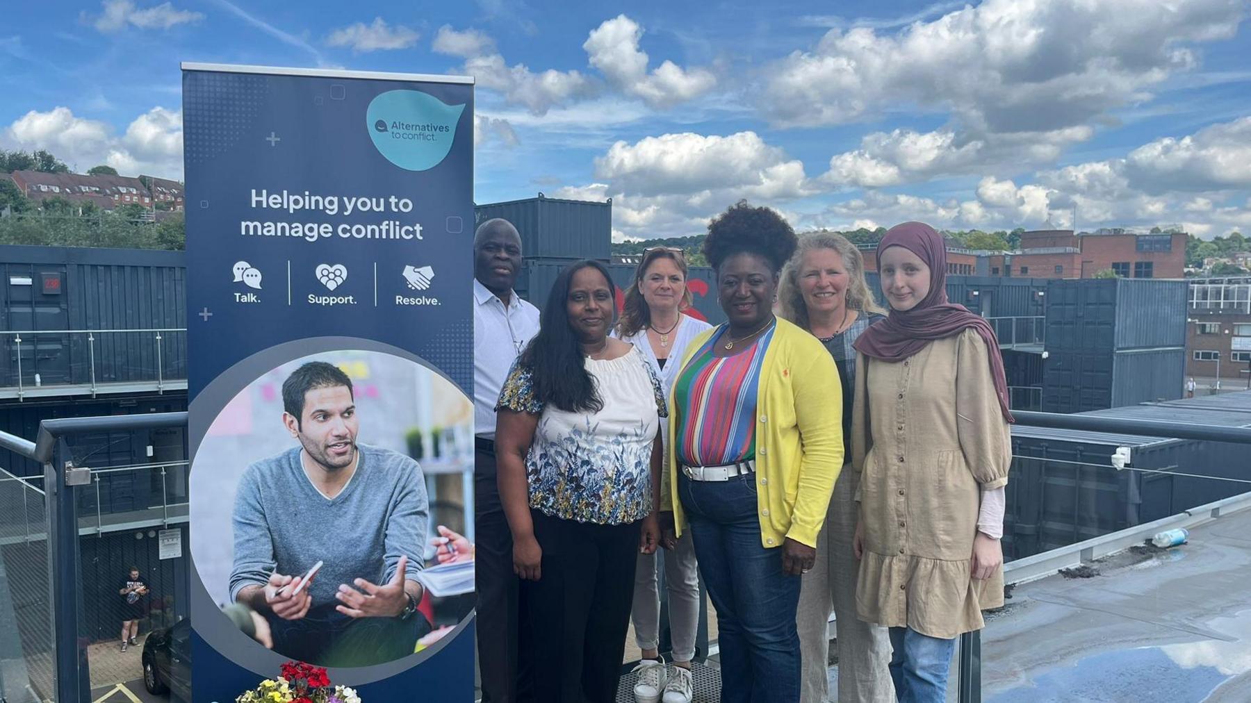 A poster with the name Alternative to Conflict on it is on the right. On the left there is a group of six people who work for the non-profit. They appear to be on a rooftop - with a skyline behind them.