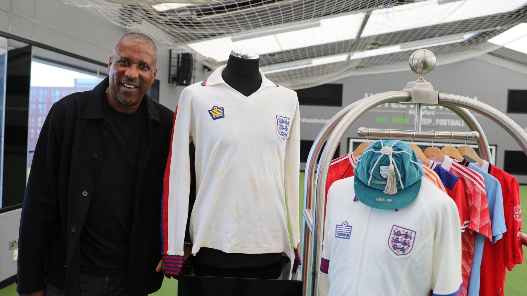 Viv Anderson, wearing a black top and jacket, stands alongside his 1978 shirt from his England debut. To the right is his tracksuit top from the same game and a blue cap awarded to players in the team. Behind, on a clothes rail, hang six other football shits, mostly red with one light blue.