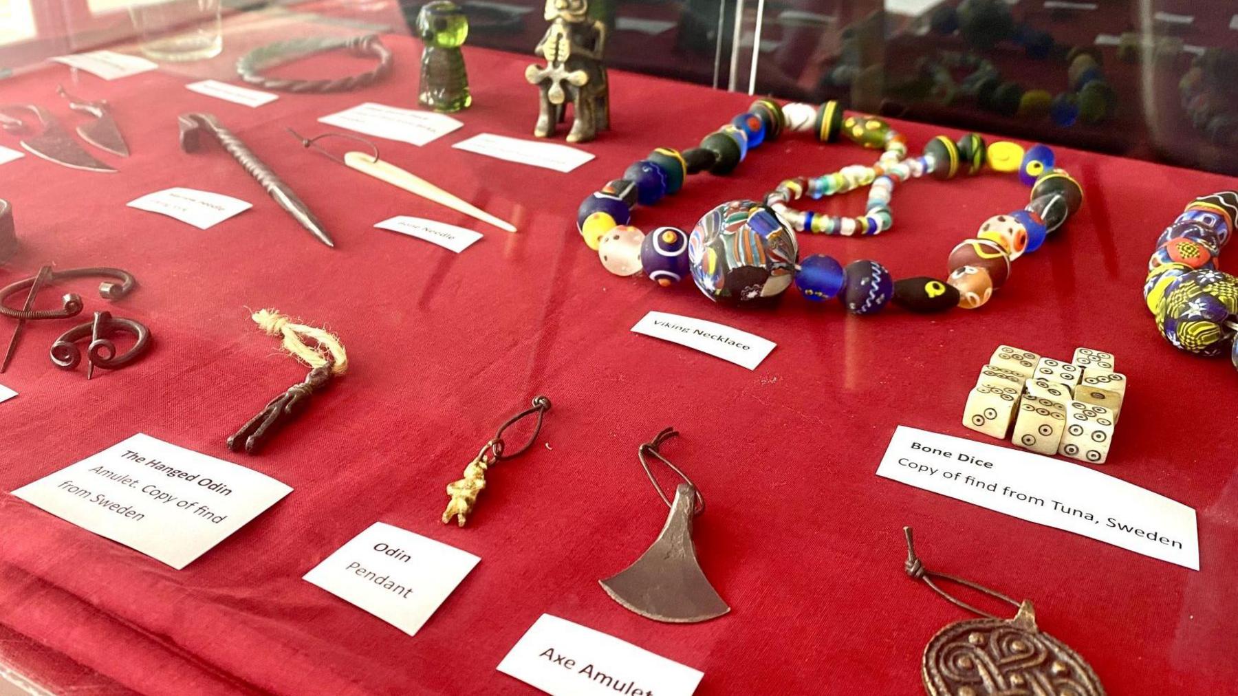 A red table with different items each has a white label beneath. The items include bone dice, an amulet and an Odin pendant.