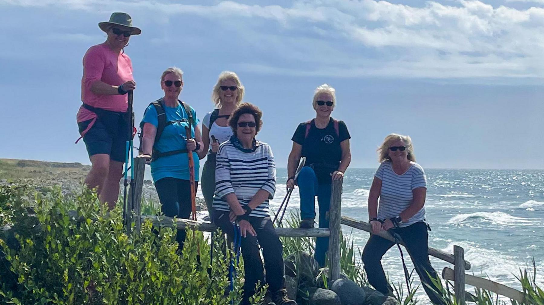 Six women from Walk Kernow stand or sit on a wooden fence during a walk between Coverack and St Keverne. It is a sunny but cloudy day. The sea is behind them.
