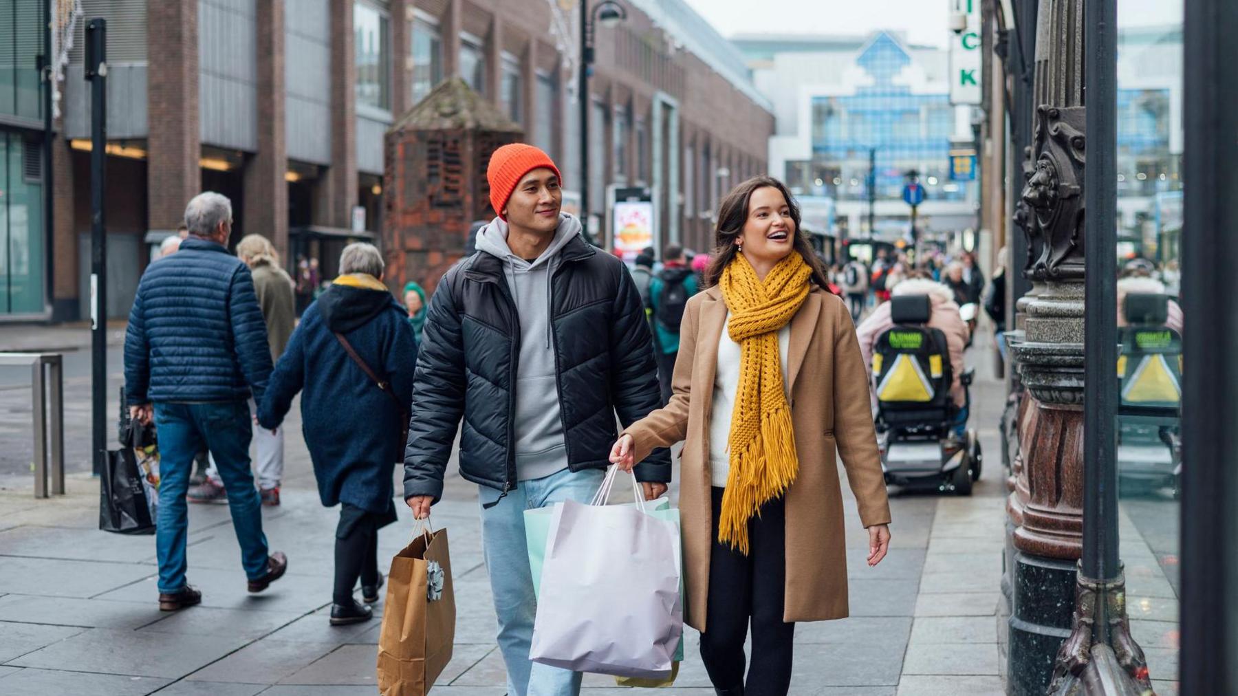 A young couple, both dressed in warm winter clothes and carrying shopping bags. They are walking on a British high street, and both are smiling.